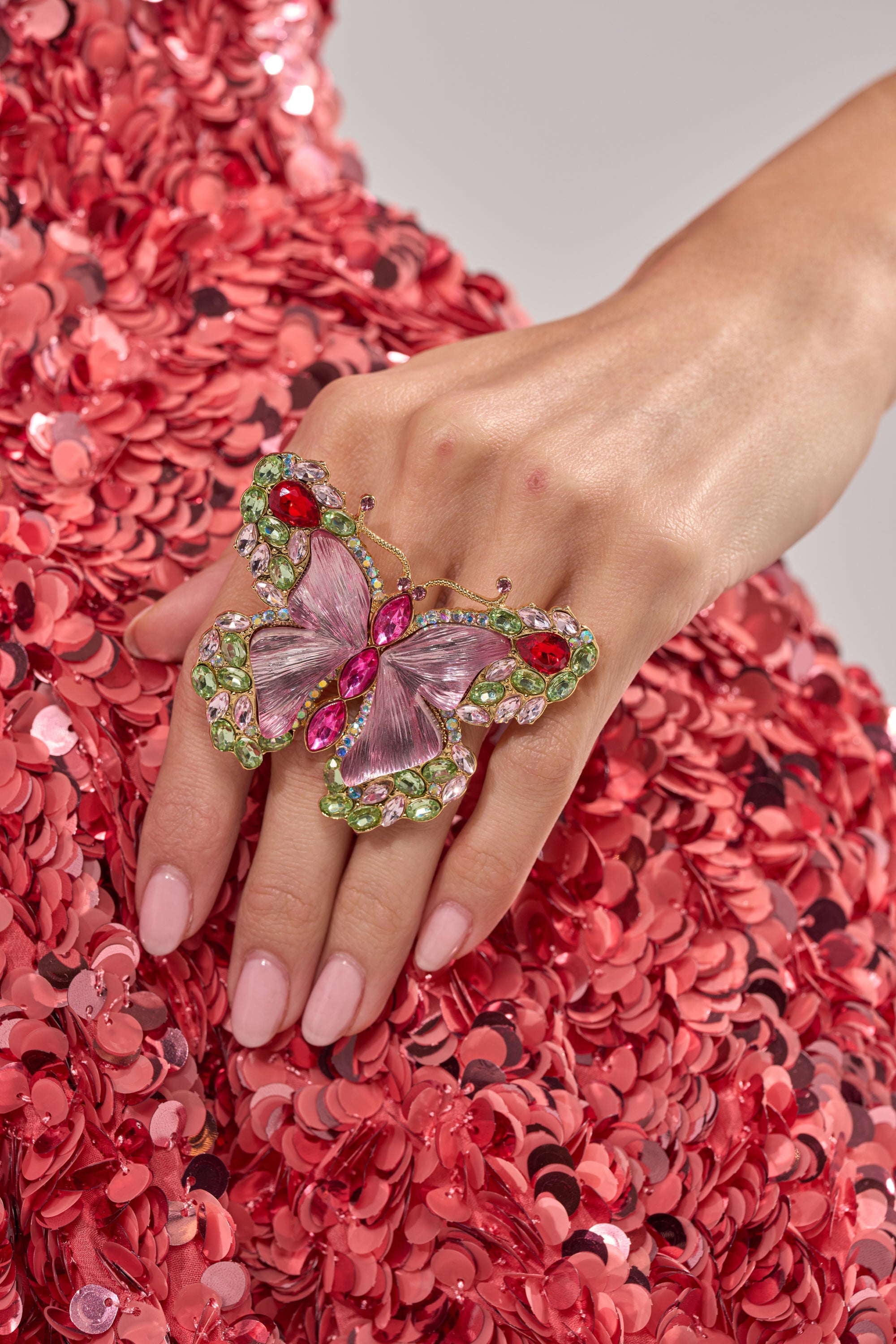 A hand with pale pink manicured nails wears the SPREAD YOUR WINGS RING, a large and colorful butterfly crystal design, set against shimmering pink sequin fabric. 7