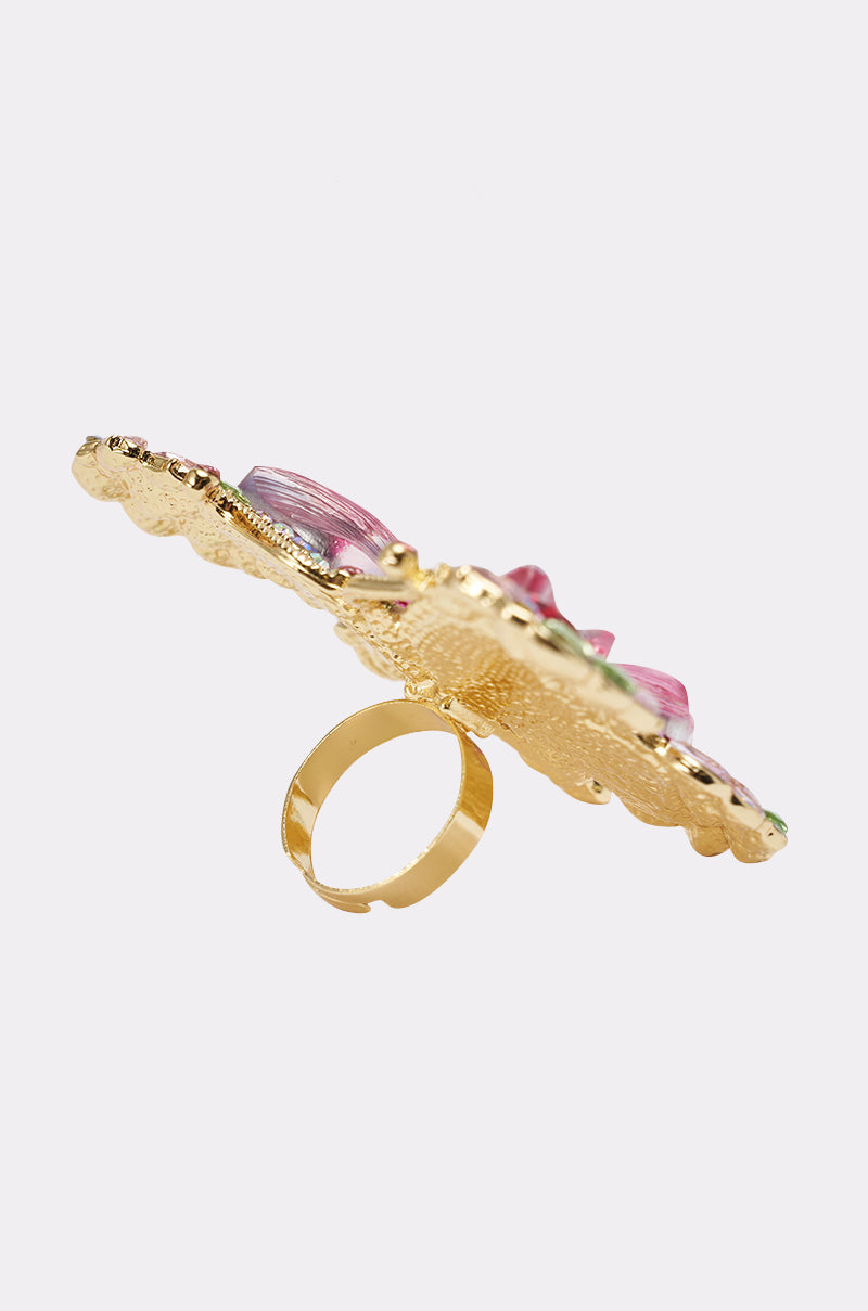 The SPREAD YOUR WINGS RING is a gold ring with a large, flat, textured oval top featuring pink and purple accents, shown from the side on a plain white background. 10