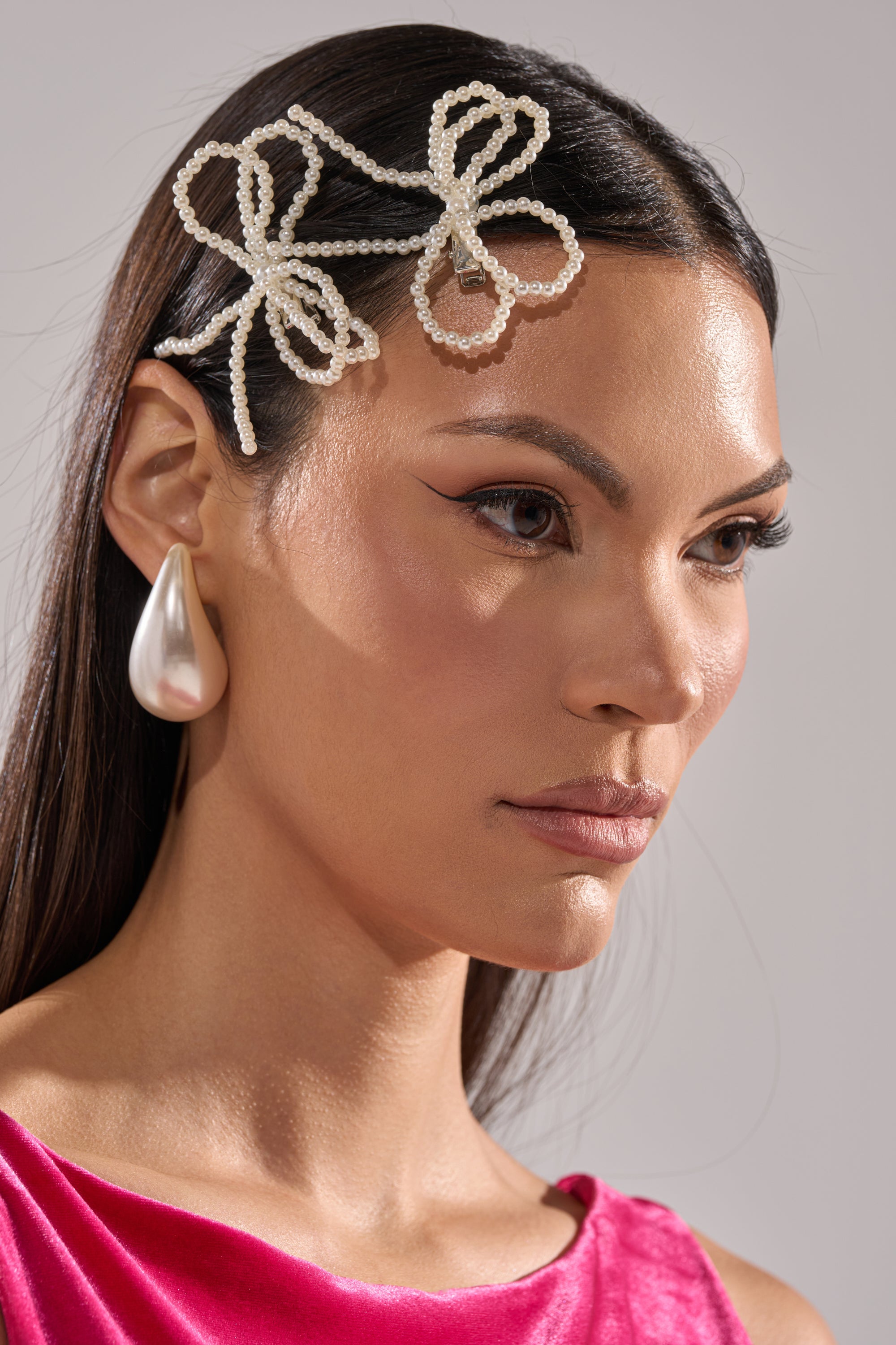 A woman with straight dark hair wears the POLLY PEARL HAIR CLIP SET, a bold pearl drop earring, subtle winged eyeliner makeup, and a shiny pink top. She gazes calmly to the side. 1