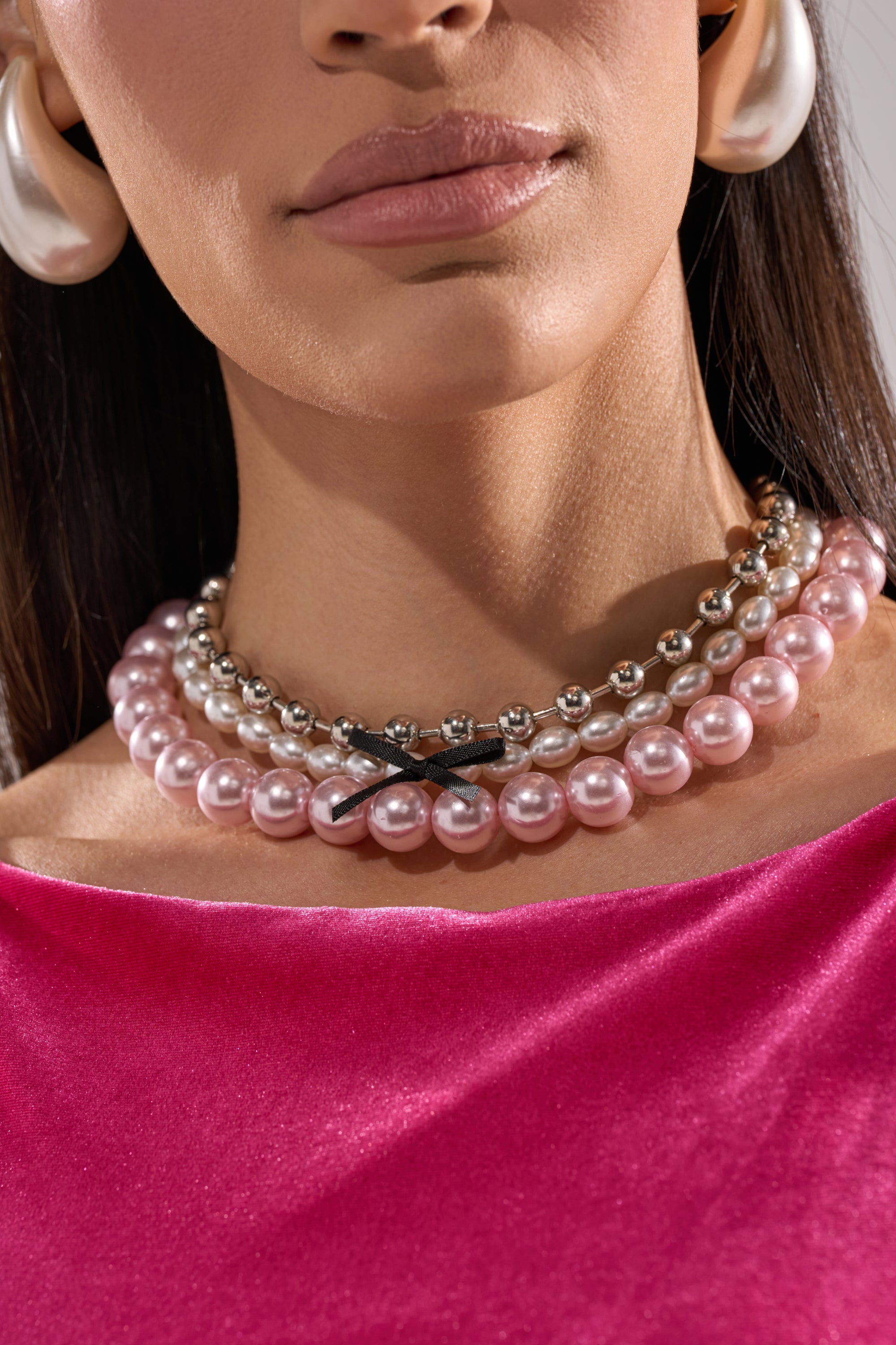 A woman showcases the GWEN NECKLACE SET featuring layered pink and silver pearls with a small black bow, paired with large pearl earrings and a bright pink velvet top. Only her lower face and neck are shown. 1
