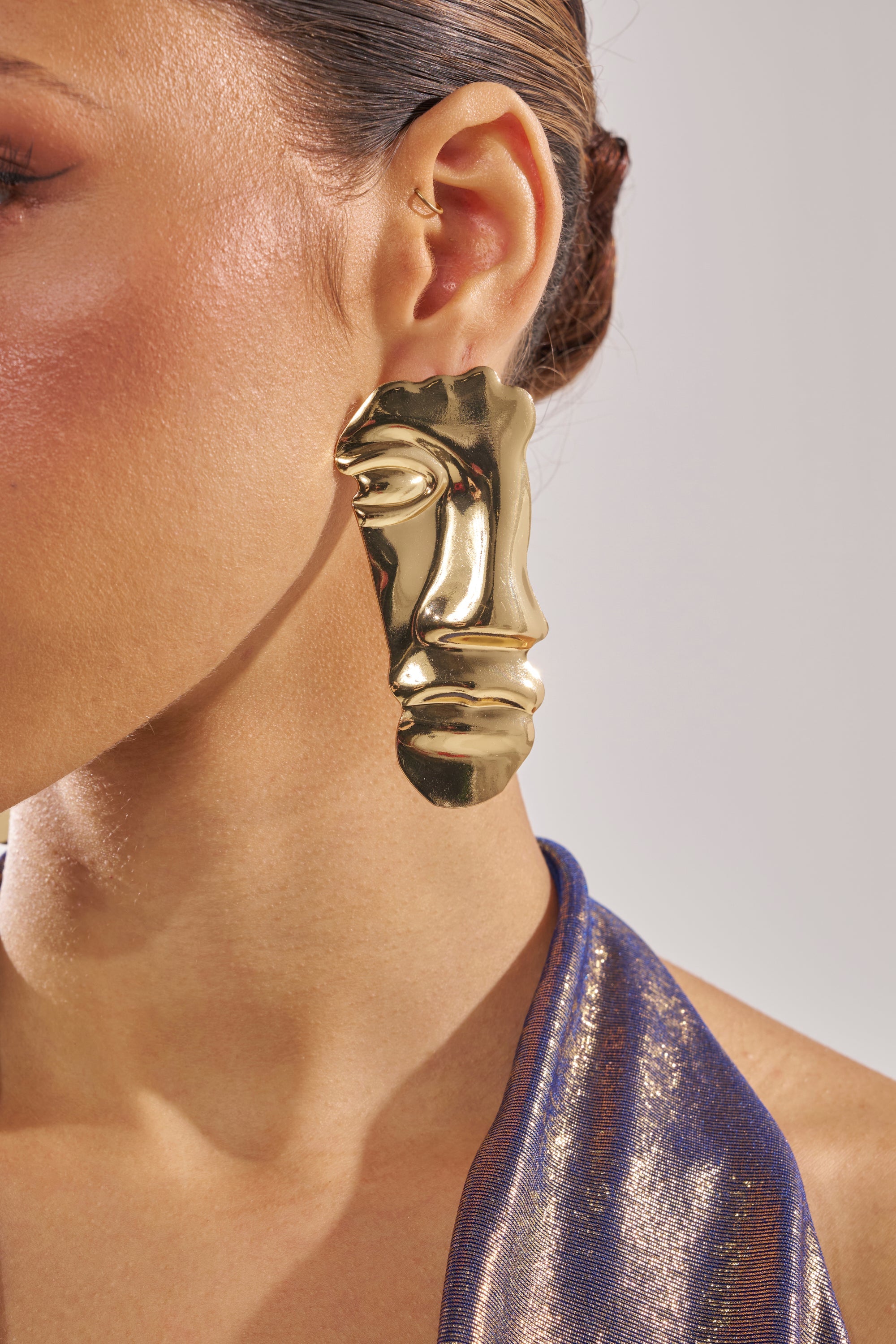 Close-up of a woman with smooth skin and hair tied back, wearing the 2FACE EARRING—gold-tone abstract earrings shaped like stylized faces—paired with a metallic blue halter top. 1