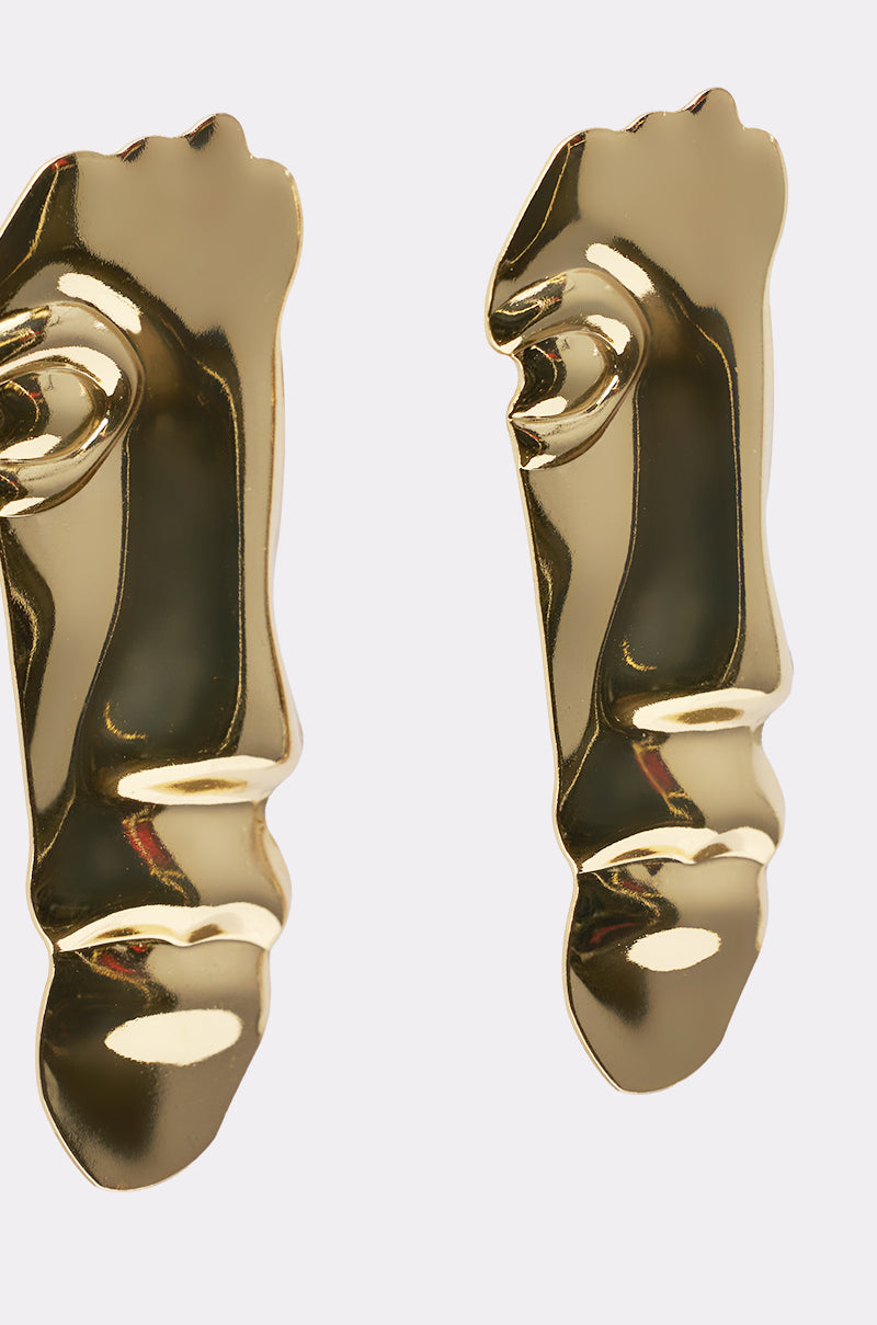 Close-up of the 2FACE EARRING: two gold-tone statement earrings showcasing stylized facial features—eye, nose, and lips—on a smooth, reflective surface. 3