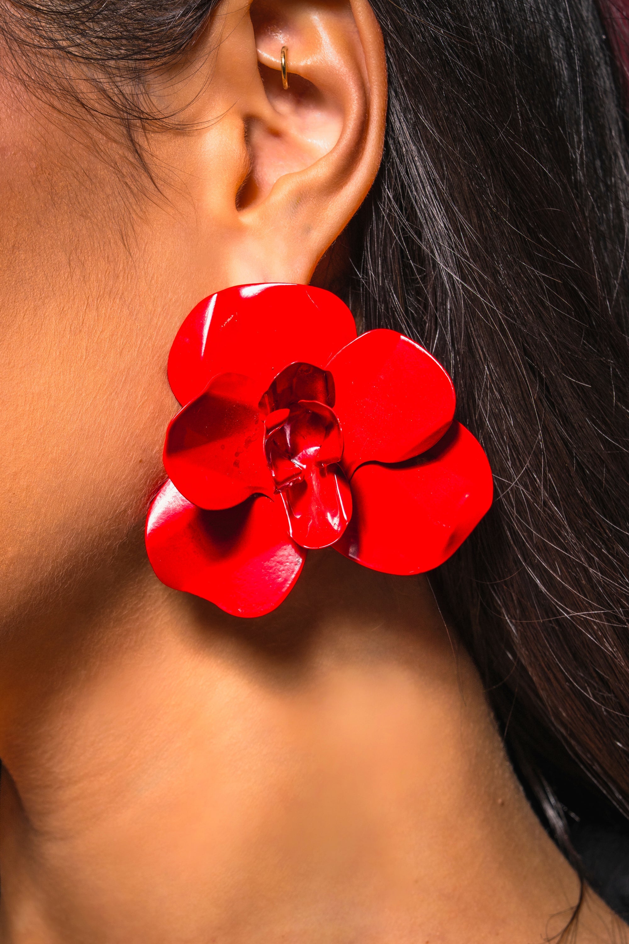 Close-up of a person wearing the GALA EARRING, a large, shiny crimson floral post earring. The person has long dark hair and also wears a small gold hoop in the upper ear. 0