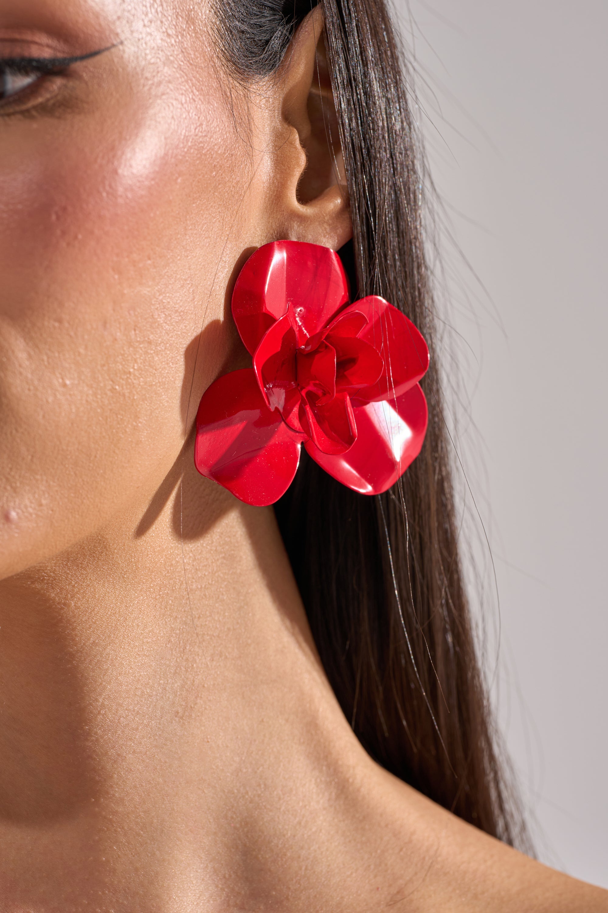 Close-up of a woman's face with smooth skin and long dark hair tucked behind her ear, showcasing the GALA EARRING—a large, shiny crimson floral post earring. 4