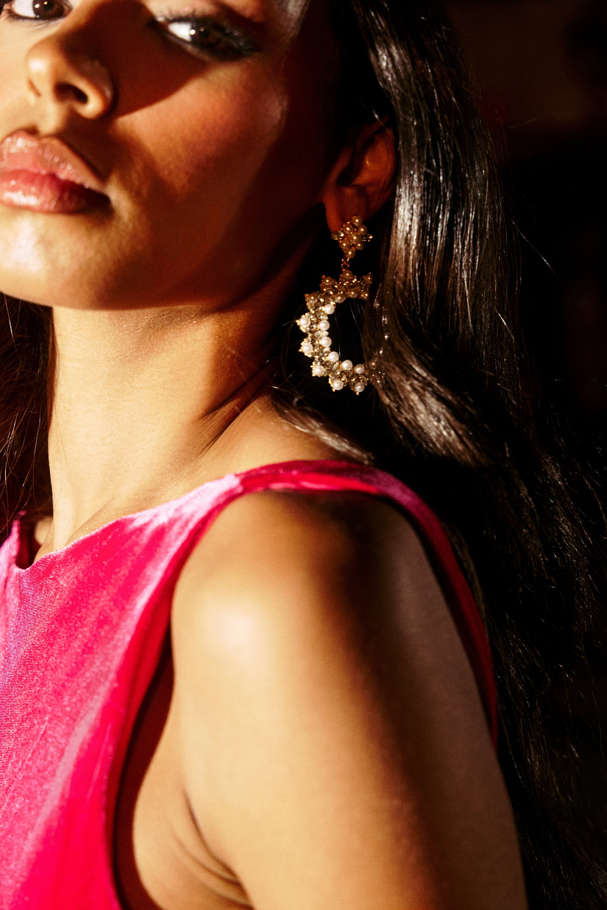 Close-up of a woman with long dark hair wearing gold hoop PEARL PLS EARRING, decorated with pearls and rhinestones, paired with a bright pink sleeveless top. Warm light highlights her face, shoulder, and sparkling jewelry.