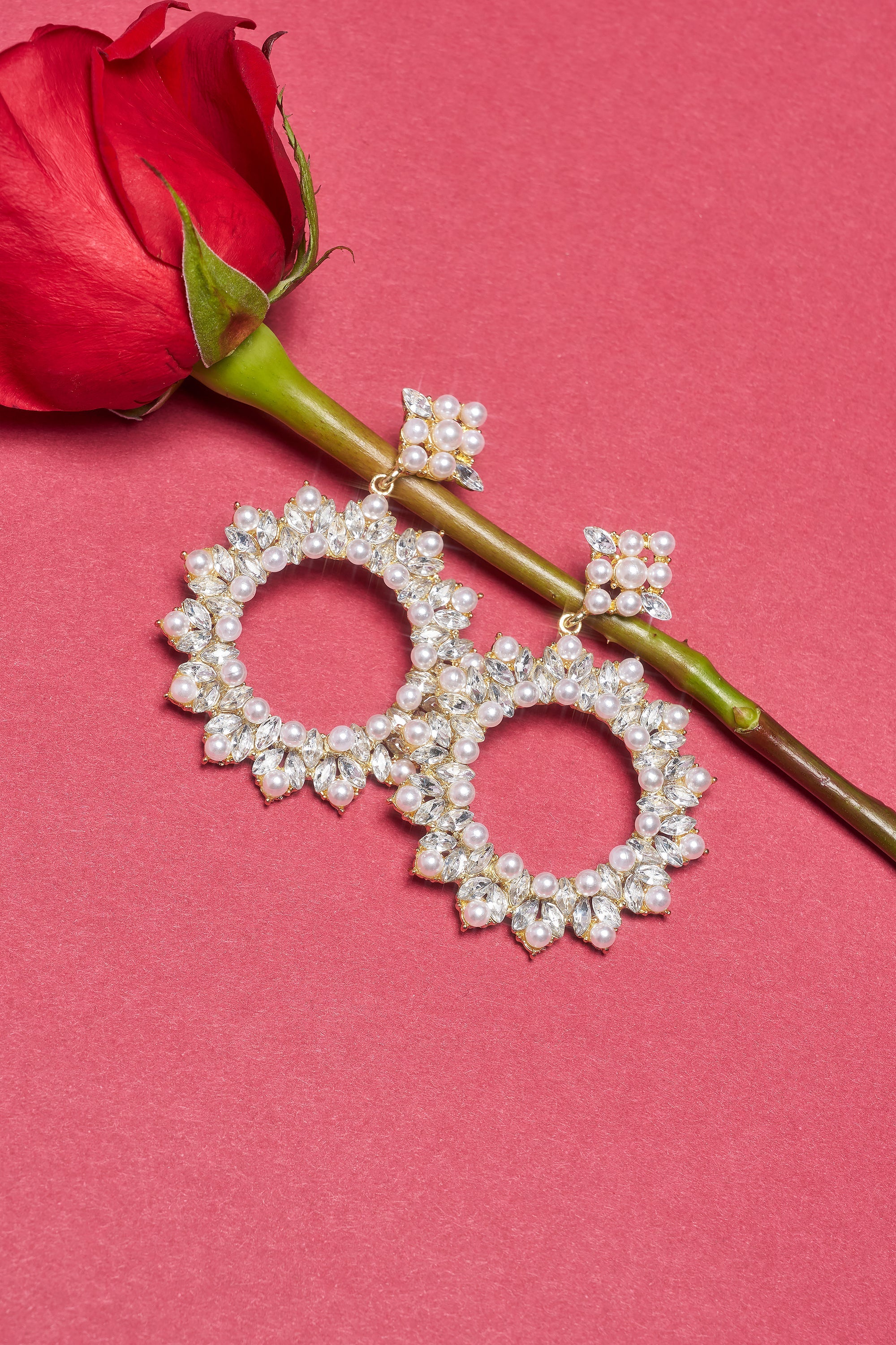 The PEARL PLS EARRING—ornate gold hoops with pearls and rhinestone trim—is displayed on a red background beside a single red rose. 0