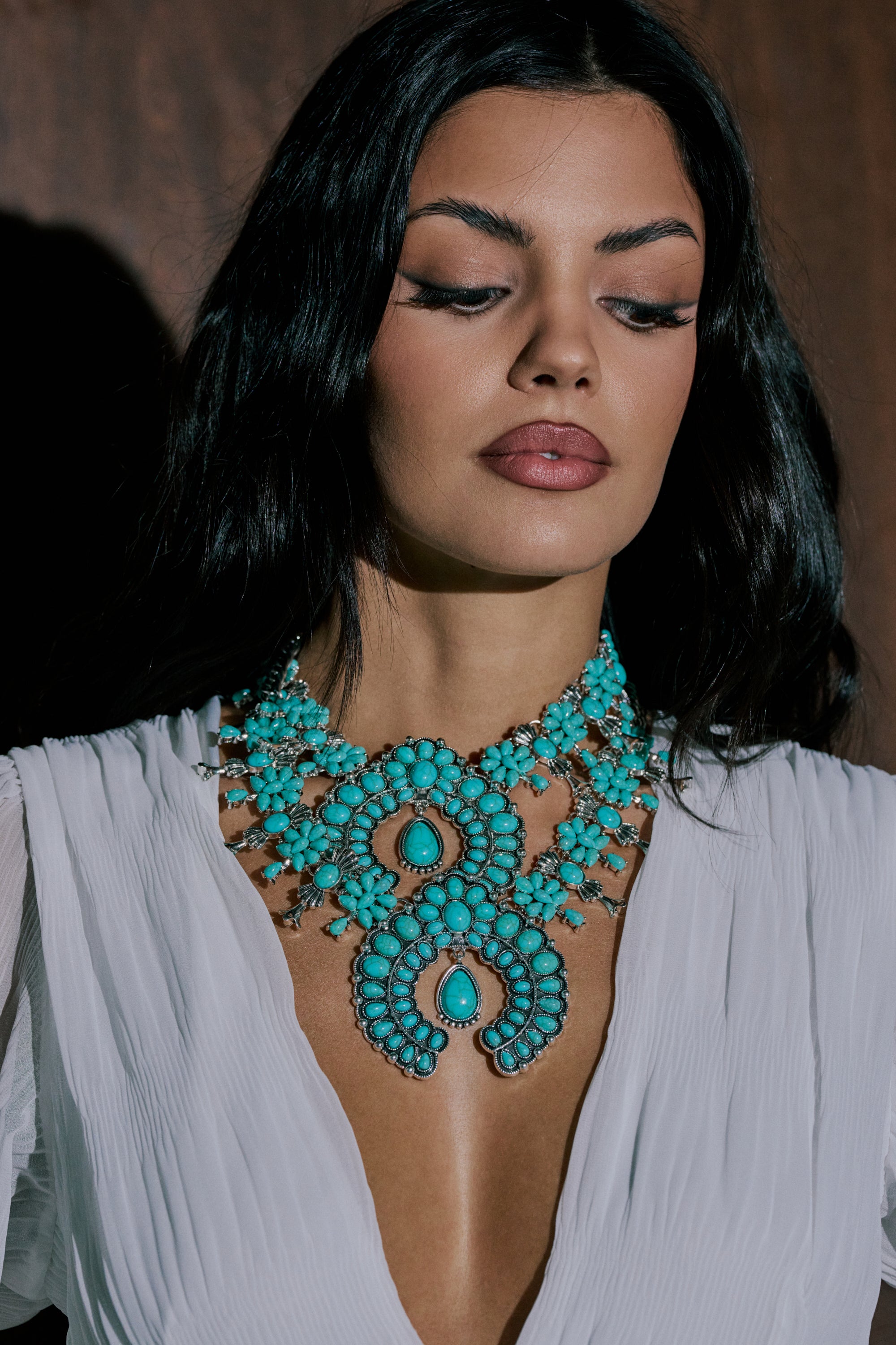 A woman with long dark hair wears a white v-neck blouse and the HERE FOR THE VIBES NECKLACE, gazing down slightly with a neutral expression against a dark background. 0