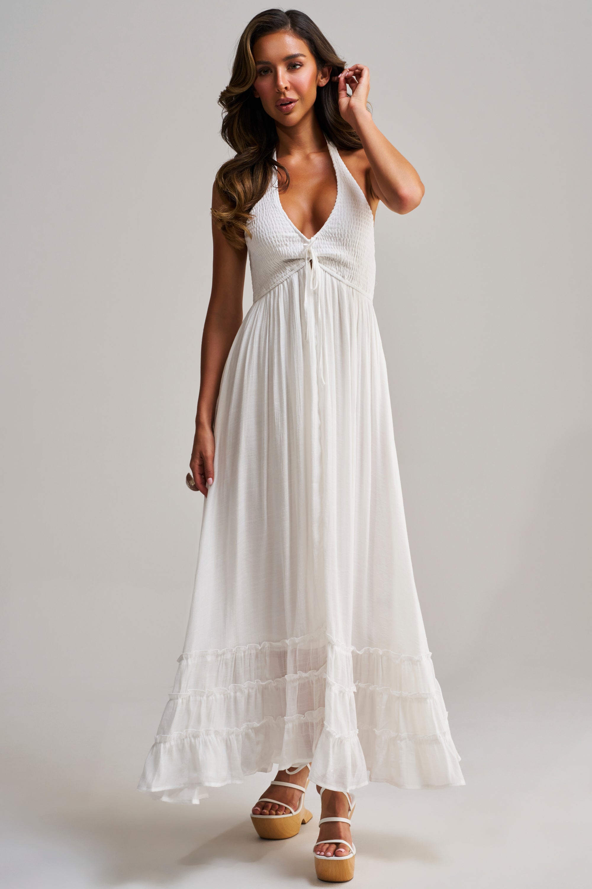 A woman stands against a plain background, touching her hair and wearing platform sandals, dressed in the THIS IS MY DATE MAXI DRESS IN WHITE—a sleeveless halter maxi with a deep V-neckline and ruffle details.