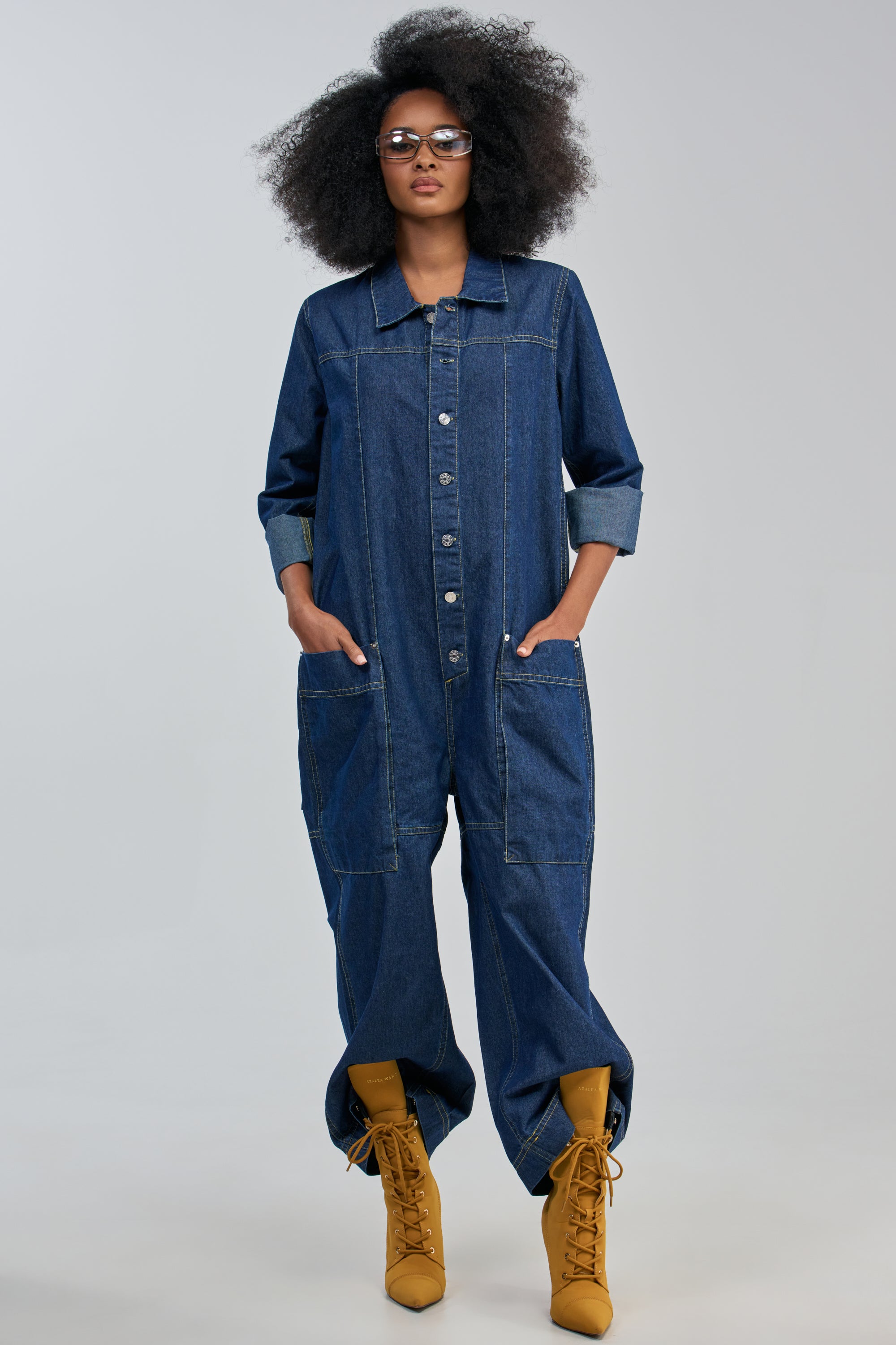 Wearing the PLANNING ON YOU LONG SLEEVE JUMPSUIT IN DARK DENIM with rolled-up sleeves and large front pockets, styled with yellow lace-up boots and glasses, this person stands hands in pockets against a plain background, effortlessly embodying streetwear jumpsuit style.