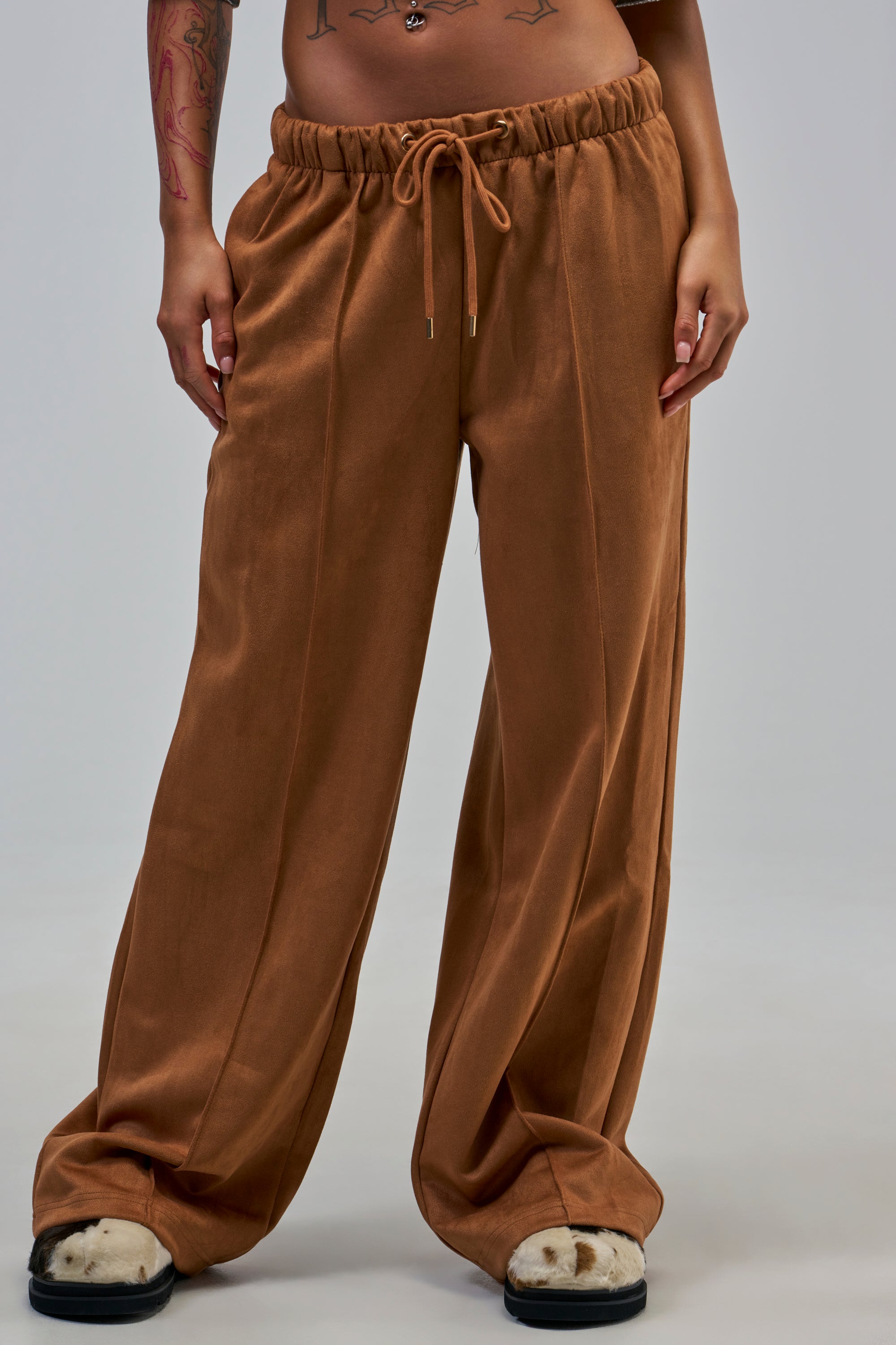 Lower torso and legs of a person wearing SHAKE IT UP SUEDE TROUSER IN BROWN—loose, wide-leg silhouette—paired with black-and-white slippers, posed against a plain background.