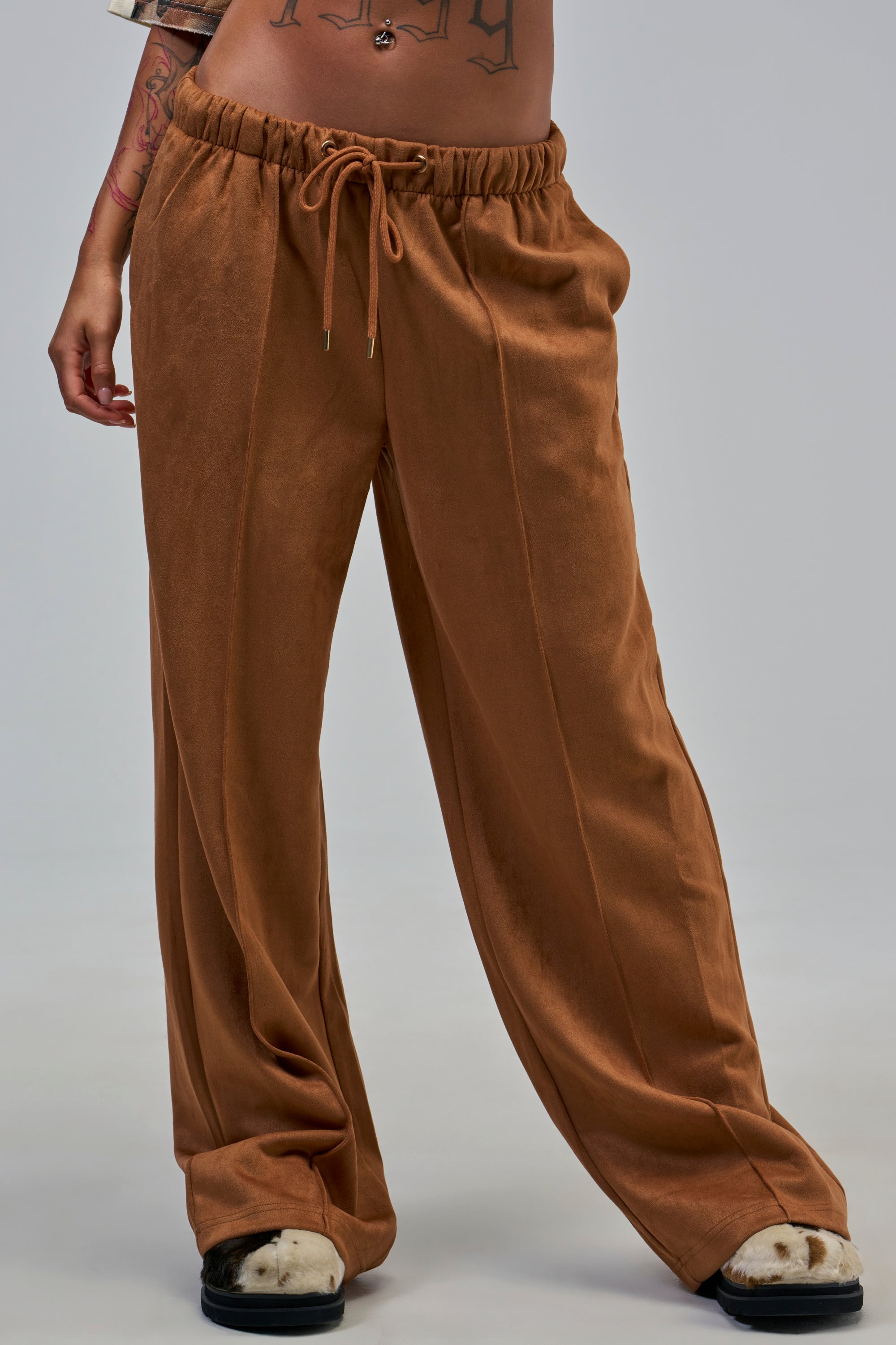 Model showcasing the SHAKE IT UP SUEDE TROUSER IN BROWN, featuring a loose, wide-leg silhouette and drawstring waist. Only the lower torso and legs are visible against a plain gray background.