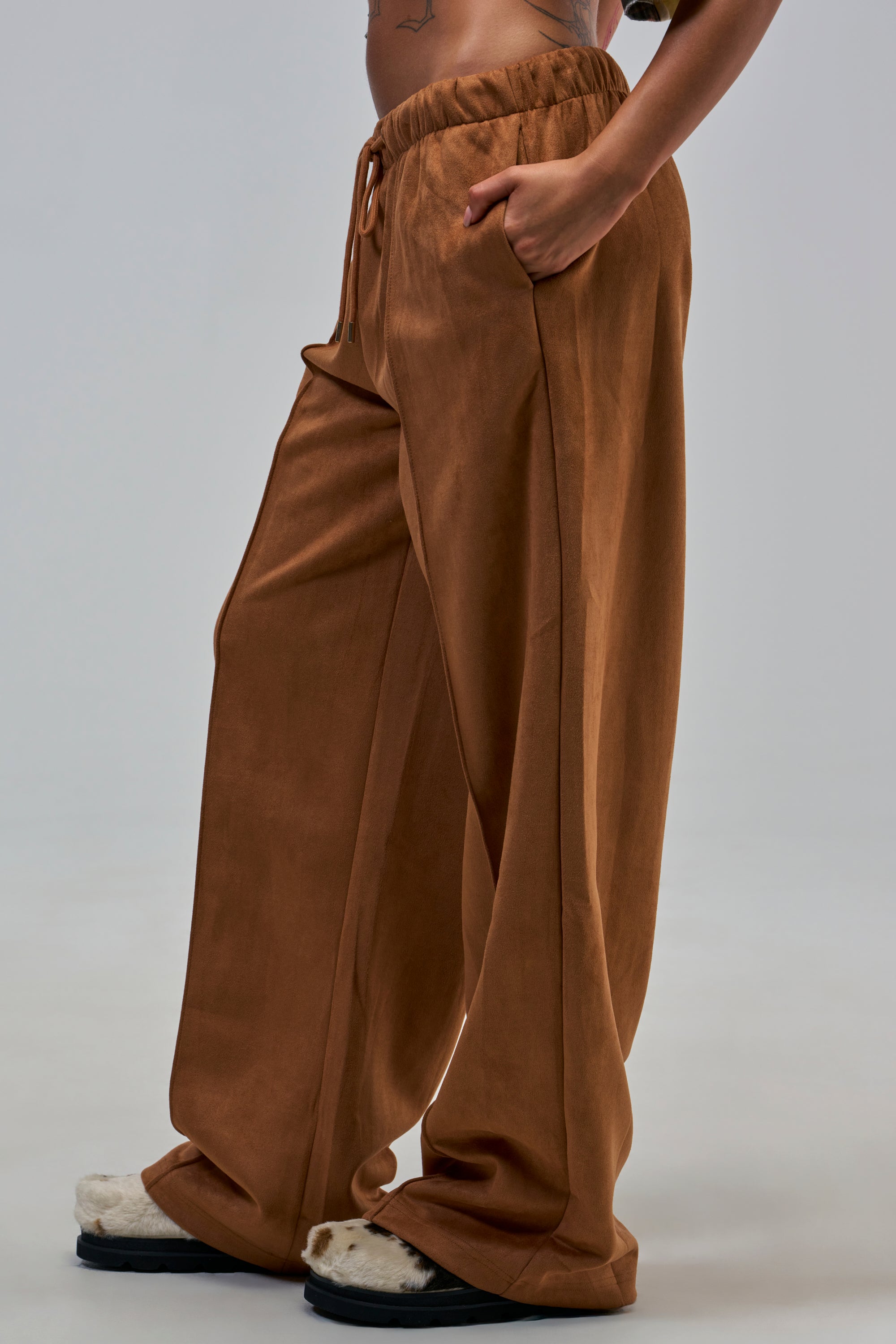A person wearing SHAKE IT UP SUEDE TROUSER IN BROWN with a wide-leg fit and fuzzy slippers stands with one hand in their pocket against a plain background.