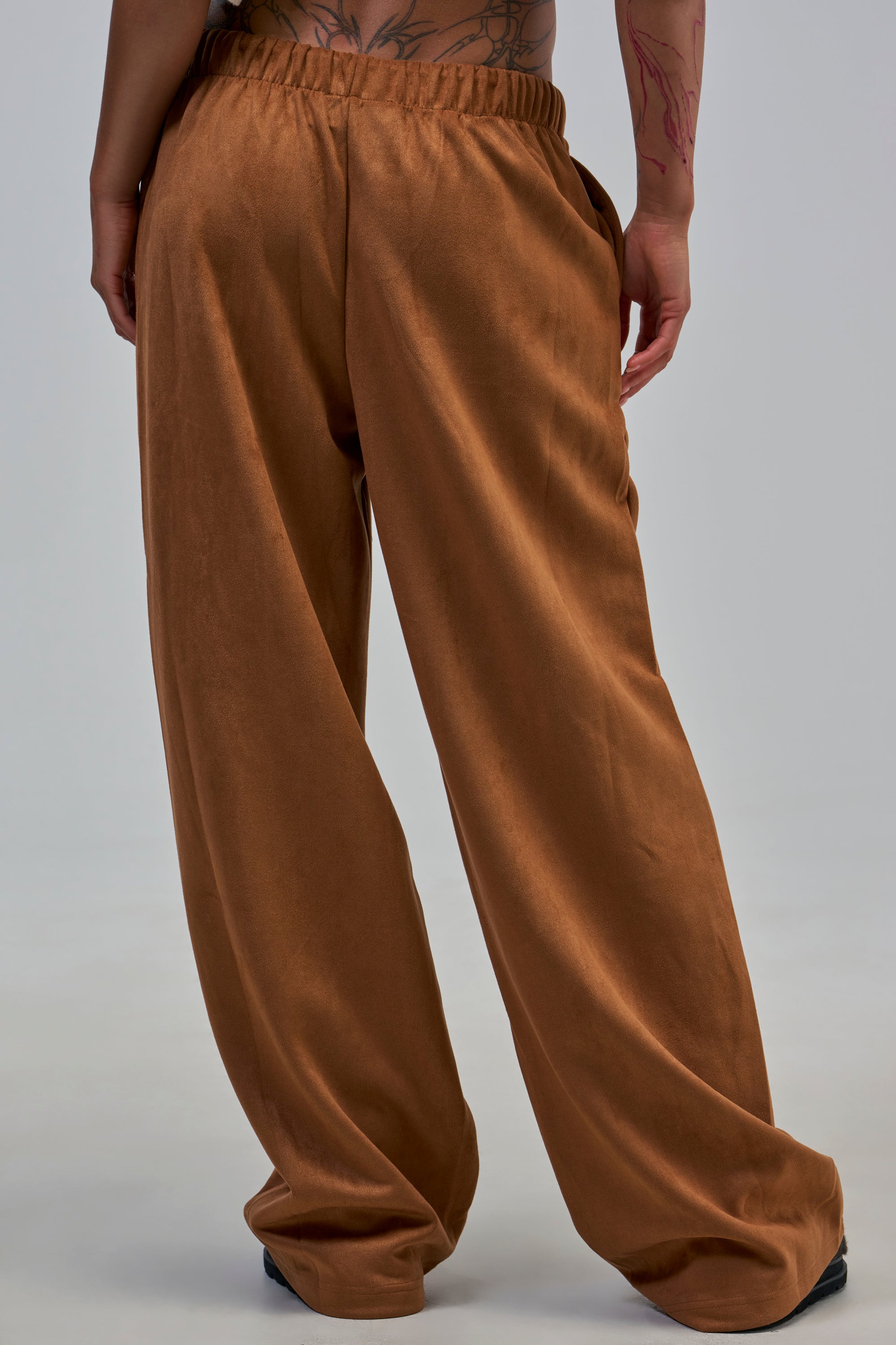 Model wearing SHAKE IT UP SUEDE TROUSER IN BROWN—loose-fitting, high-rise suede trousers—with black shoes, back to camera; tattoos visible on left arm against light gray background.