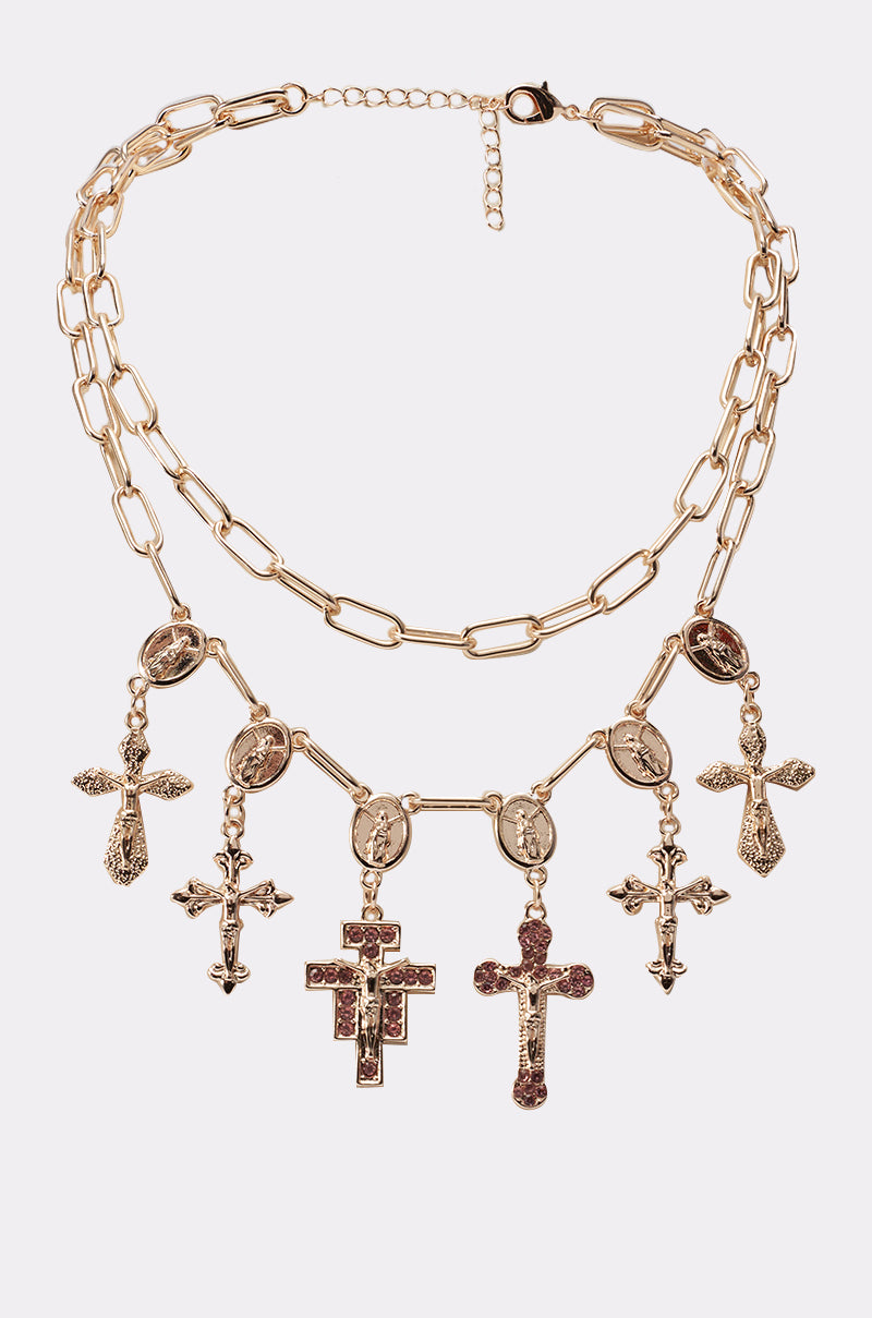 The PRETTY IN PINK NECKLACE is a gorgeous gold layered piece with two chains, featuring five ornate cross and angel pendants. Intricate lower chain charms are accented with red gemstones for a striking effect. 1