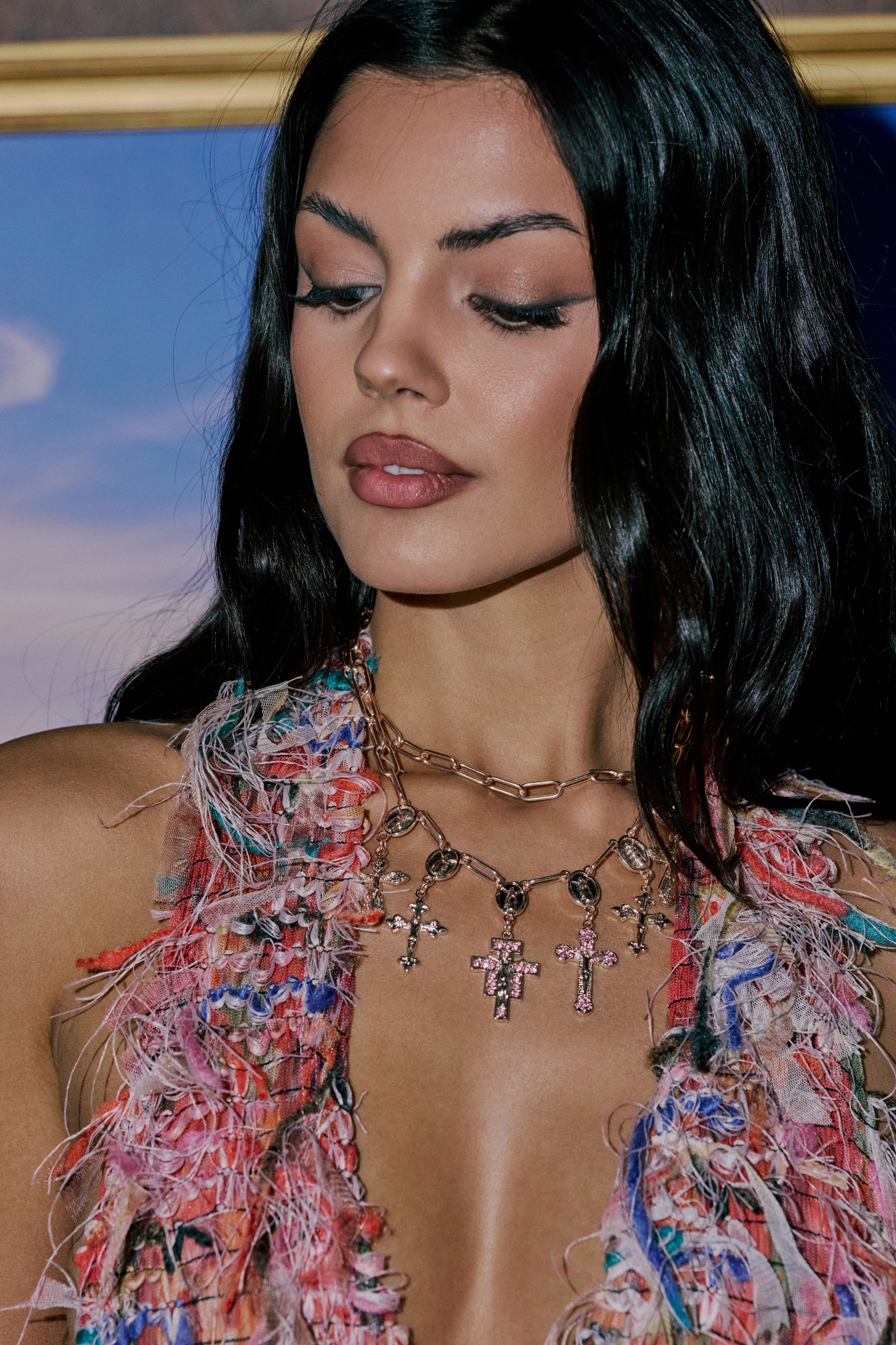 A woman with long dark hair wears a colorful fringed top and the PRETTY IN PINK NECKLACE, its shimmering charms catching the light as she gazes downward with a calm expression. 0