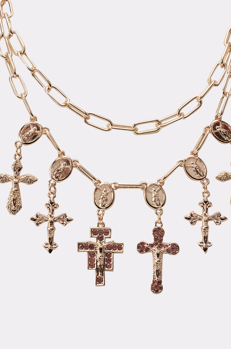The PRETTY IN PINK NECKLACE is a layered gold chain adorned with multiple religious charms, such as cross pendants and oval saint medallions, displayed against a plain white background. 2