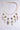 The JEAN NECKLACE features gold-tone links with large chain links, accented by multiple decorative pendants like crosses and ornate designs, elegantly showcased against a light gray background.