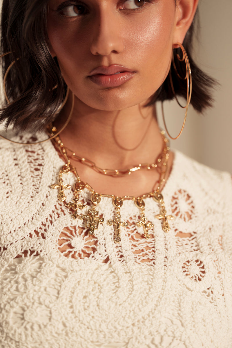 A woman from the shoulders up wears a white crocheted top, large gold hoop earrings, and the JEAN NECKLACE featuring double chains with cross pendants. The softly lit image highlights her accessories.