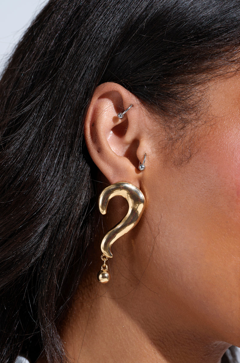 A close-up of a person's ear with three piercings displays the eye-catching EXCUSE ME EARRING, featuring a large gold question mark with a hammered-metal finish that pairs perfectly with dark hair against a plain white background. 1