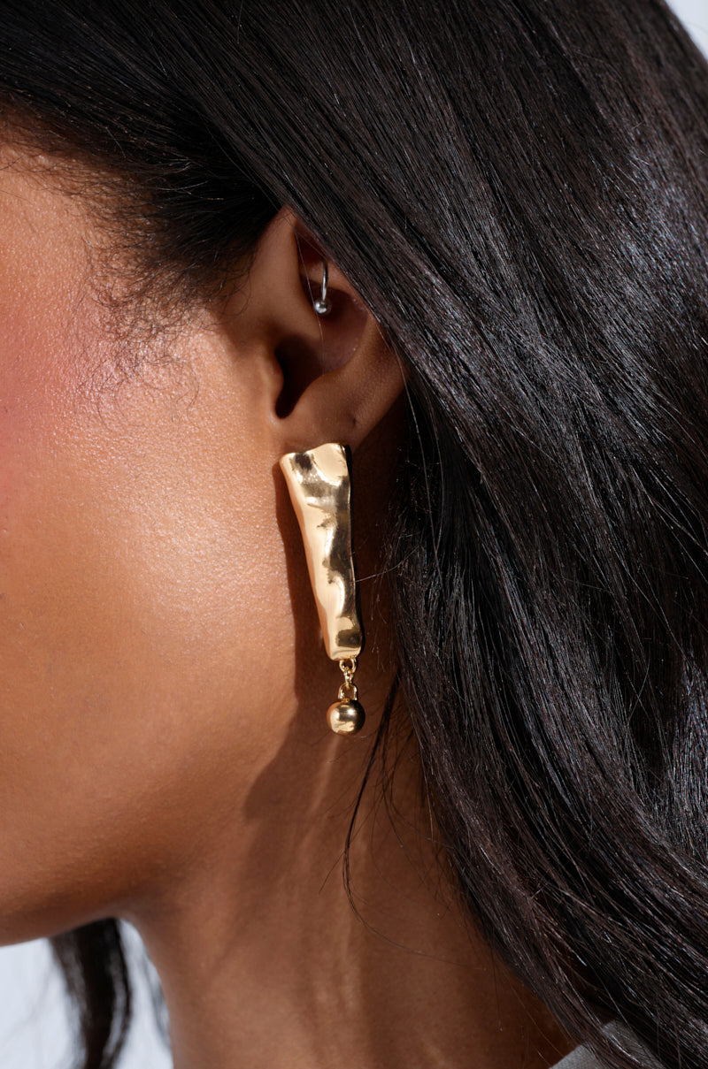 A close-up shows an ear adorned with the exquisite EXCUSE ME EARRING, a gold-tone piece featuring a hammered-metal finish in an abstract shape with a dangling sphere. A delicate hoop piercing at the top complements elegantly cascading hair. 2