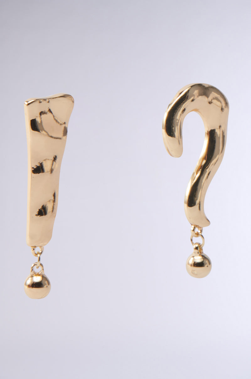 Introducing the EXCUSE ME EARRING: gold-tone earrings shaped like an exclamation mark and a question mark, each with a hammered-metal finish and a small dangling ball at the bottom, elegantly displayed on a plain gray background. 4