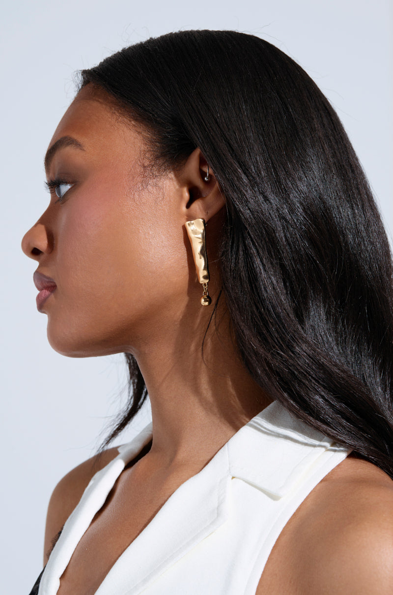 A woman with long, dark hair wears a sleeveless white top, highlighting the EXCUSE ME EARRING—a gold-tone accessory with a hammered-metal finish shaped like a paperclip. She faces left against a plain, light background. 3