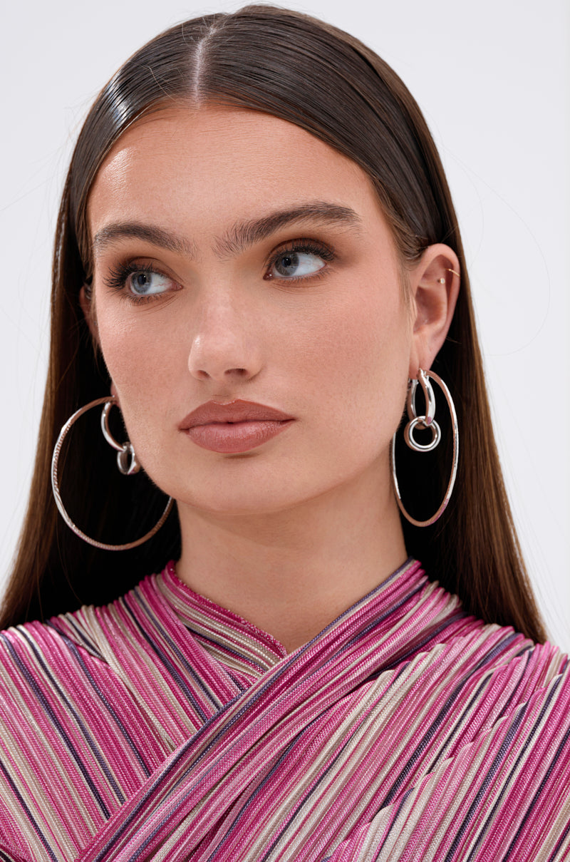 A woman with straight brown hair wears the TOO DIFFERENT HOOP IN SILVER and a pleated striped top in pink, purple, and cream shades. She gazes slightly to the side with a neutral expression. 3