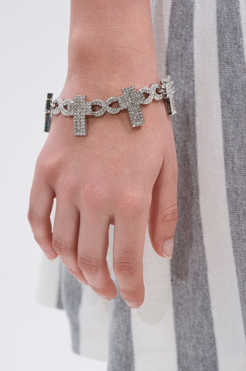A close-up of a hand wearing the CROSS MY HEART BRACELET, showcasing oversized cross pendants and rhinestone charms, set against a gray and white striped fabric background. 1