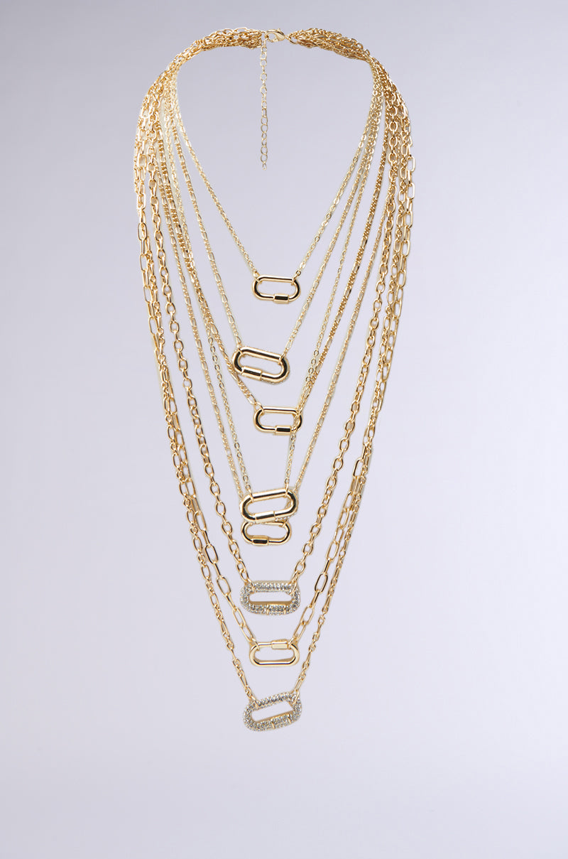 The TOO LUXE NECKLACE showcases ten symmetrically layered gold chains of varying lengths, each with a gold rectangular link. The longest chain features a rhinestone-encrusted link, creating a bold statement against the neutral gray background.