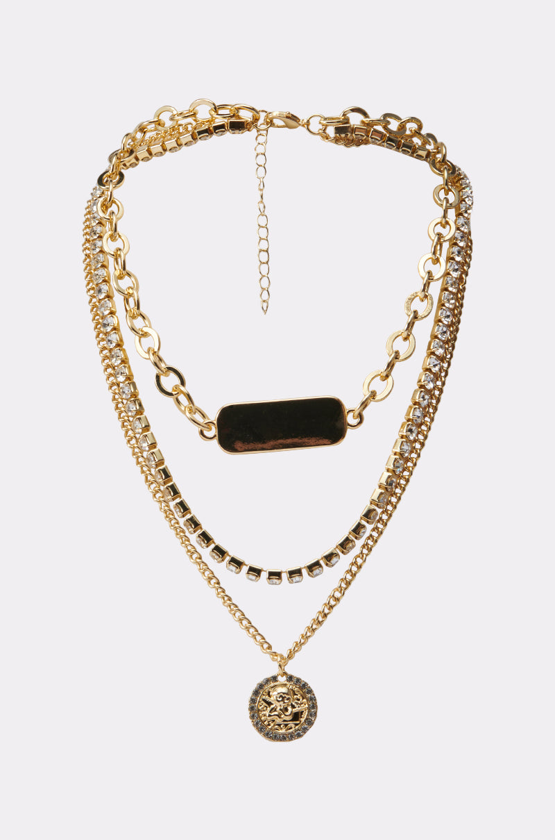 The REGENCY NECKLACE IN GOLD features three unique gold chains: a chunky link, one with clear rhinestones, and another with a lion medallion pendant, all beautifully displayed on a white background. 1