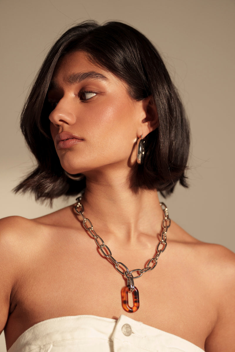 A woman with short dark hair wears a white strapless top and the MIXED FEELINGS NECKLACE, a chunky chain-link necklace with a large tortoise shell pendant and matching hoop earrings, looking to the side against a neutral background.