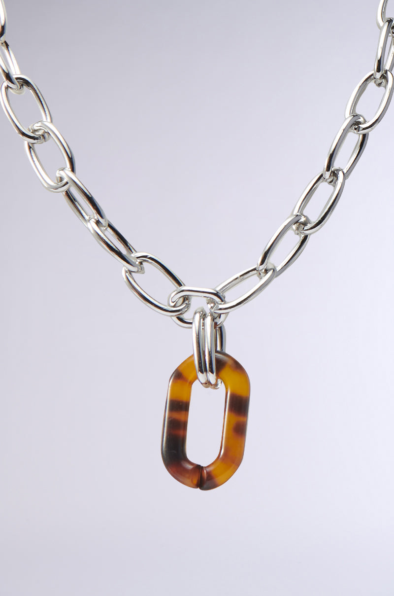 The MIXED FEELINGS NECKLACE features a chunky faux silver chain with elongated links and an eye-catching oval tortoise shell pendant, prominently displayed against a plain white background.