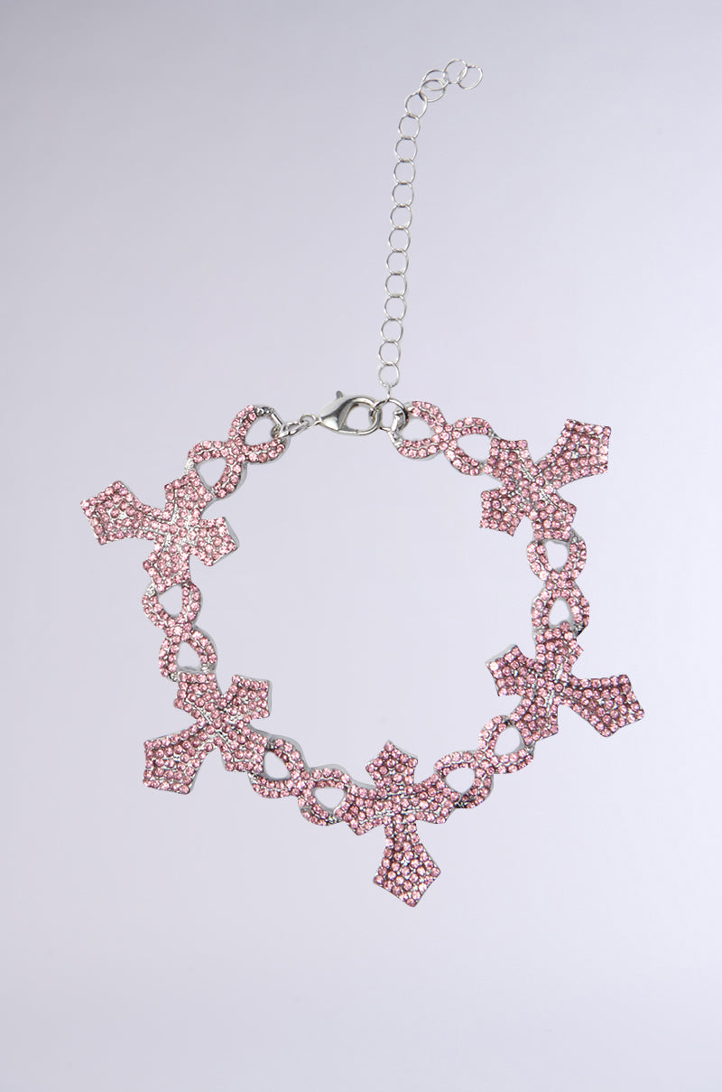The CROSSED OUT BRACELET is a silver chain featuring infinity hardware, multiple pink rhinestone cross charms, and an adjustable extender, styled against a light gray background.