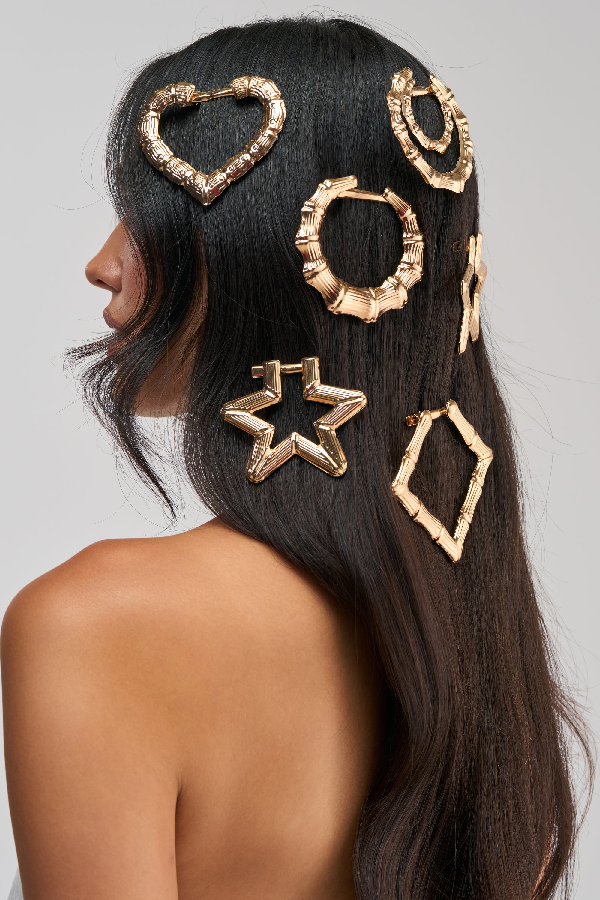 A woman with long dark hair wears the BOLD GLAMOUR HAIR CLIP SET, which features large gold alligator clips shaped like a heart, hexagon, circle, star, and diamond, elegantly arranged in her hair. She is turned to the side with her back visible against a neutral background.
