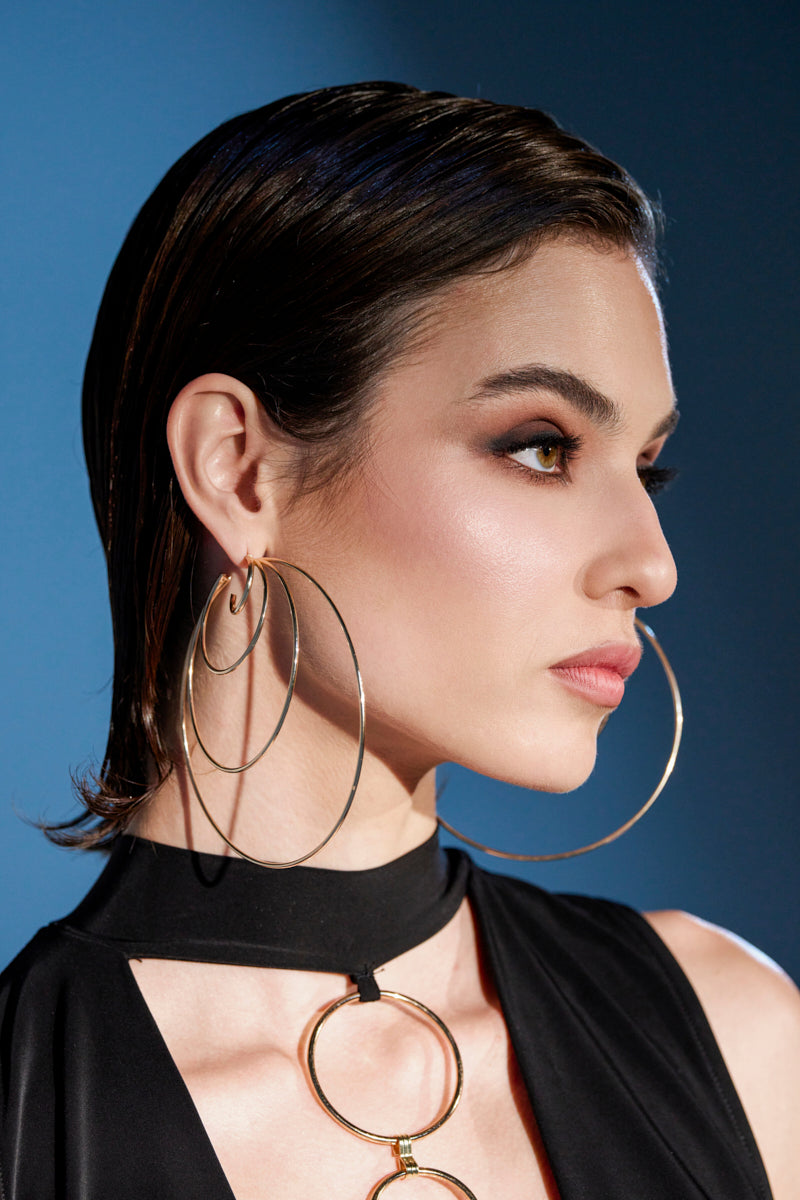 A woman with slicked-back dark hair wears the CANT KEEP UP HOOP EARRING—large faux gold hoops—paired with a black outfit with gold accents. Her bold makeup features smoky eyeshadow and highlighted cheekbones, set against a blue background.