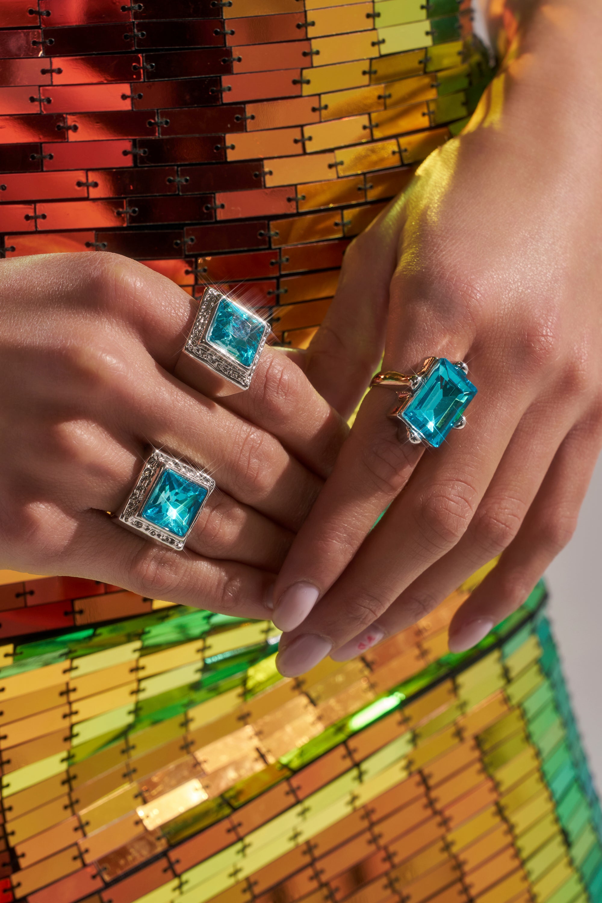 Close-up of manicured hands wearing the OUTTA THE BOX RING SET, featuring geometric rings with large square-cut blue gemstones, displayed against a vibrant sequin outfit in orange, yellow, and green tones.