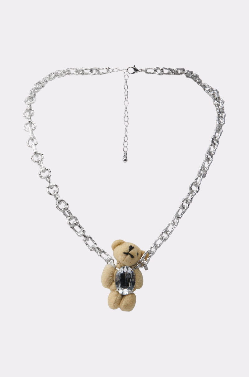 The HOLD ME TIGHT NECKLACE is a silver-tone chain with a beige teddy bear plush pendant featuring a large clear gemstone on its chest, shown against a plain white background.