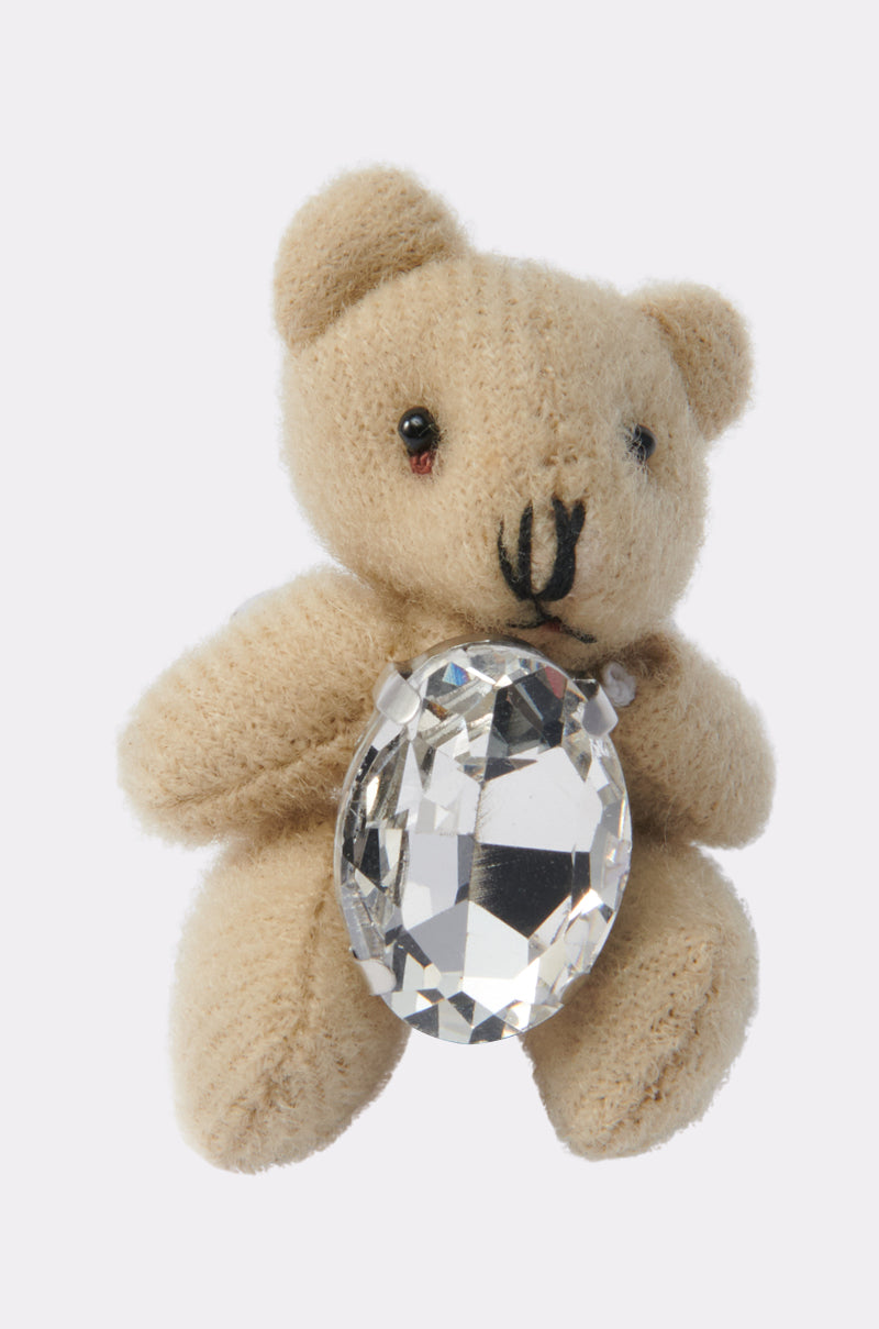 A small beige teddy bear holds the HOLD ME TIGHT RING, a large oval rhinestone piece, in its arms against a plain light background.