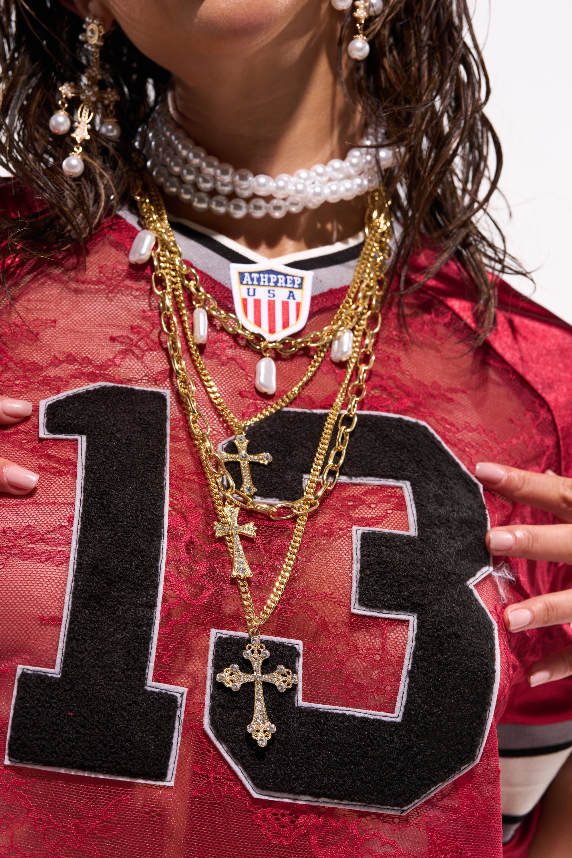A person wears the TAKE ME ON A TRIP NECKLACE—a layered chain with cross pendants—paired with pearl jewelry and a red lace top featuring a bold black 13. Damp hair frames their face as their hands rest near the necklace. 0