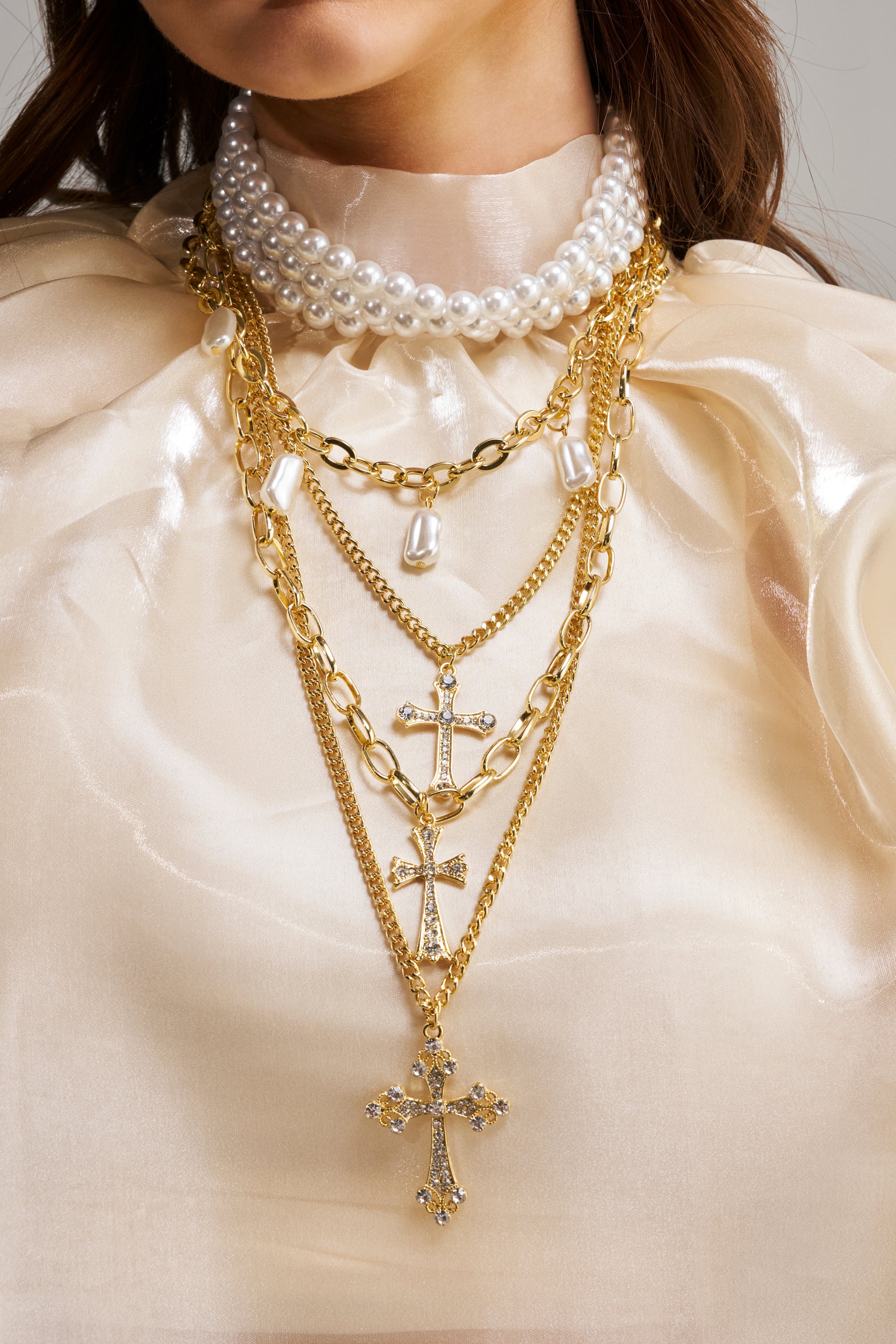 A woman wears the TAKE ME ON A TRIP NECKLACE—a layered pearl and gold chain set with a cross pendant—over a cream satin blouse, styled with festival-inspired jewelry for an elegant look.