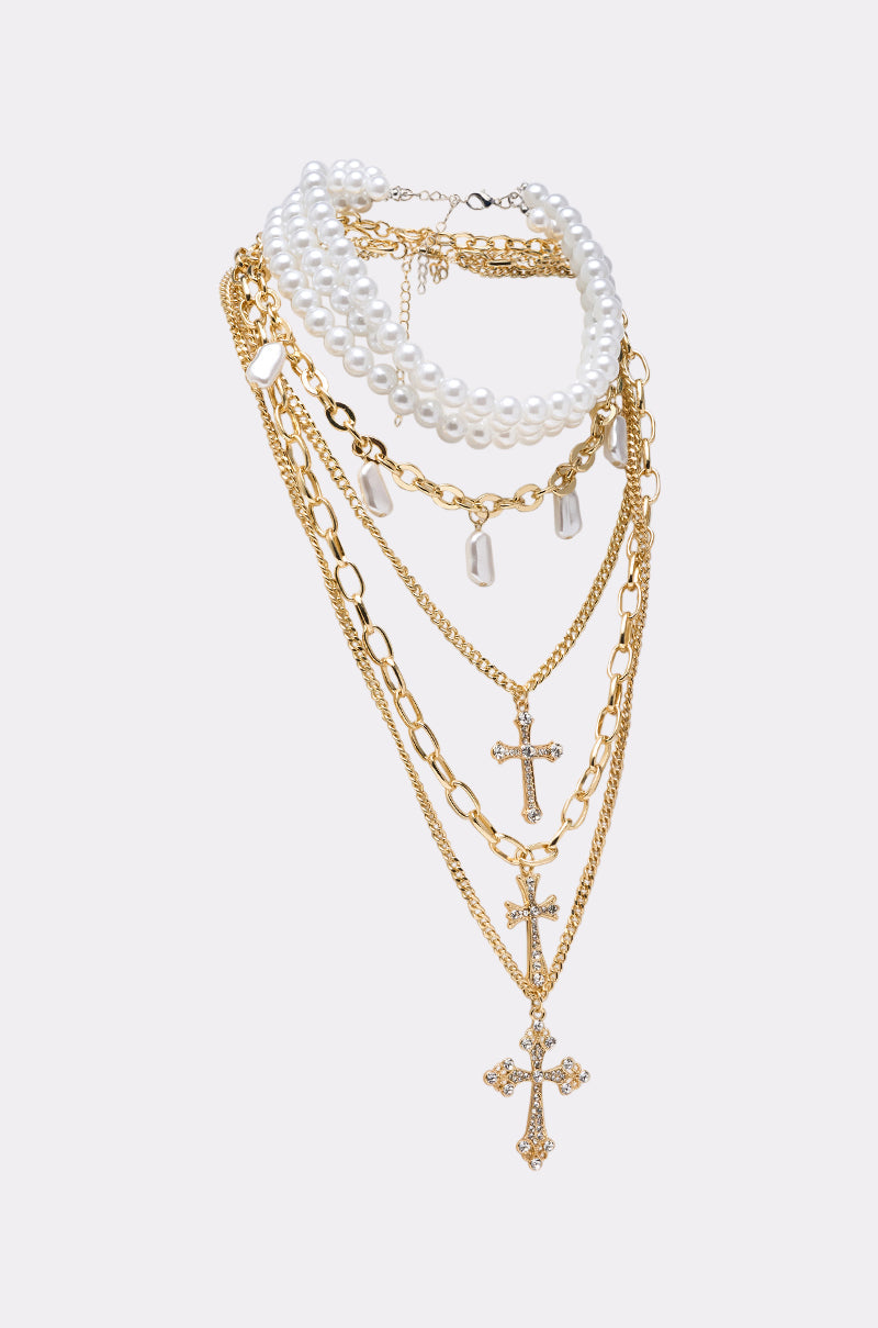The TAKE ME ON A TRIP NECKLACE features layered gold chains, a cross pendant, pearl strands, and pearl-like charms, all elegantly arranged on a white background. 3