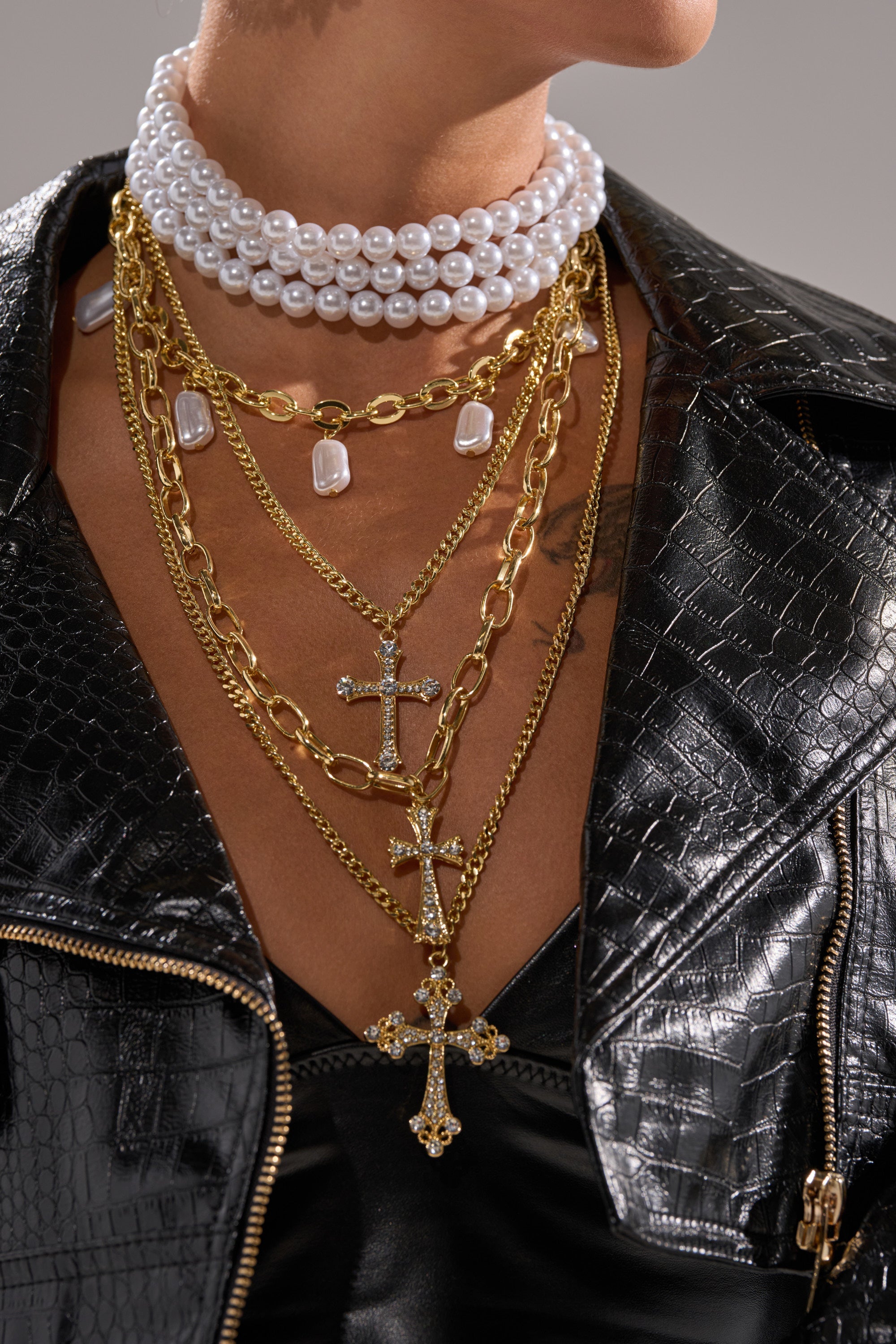 A person wears the TAKE ME ON A TRIP NECKLACE, featuring layered chains with cross pendants and a pearl choker, styled with a black faux-crocodile leather jacket over a black top. The photo highlights their neck and upper chest. 4