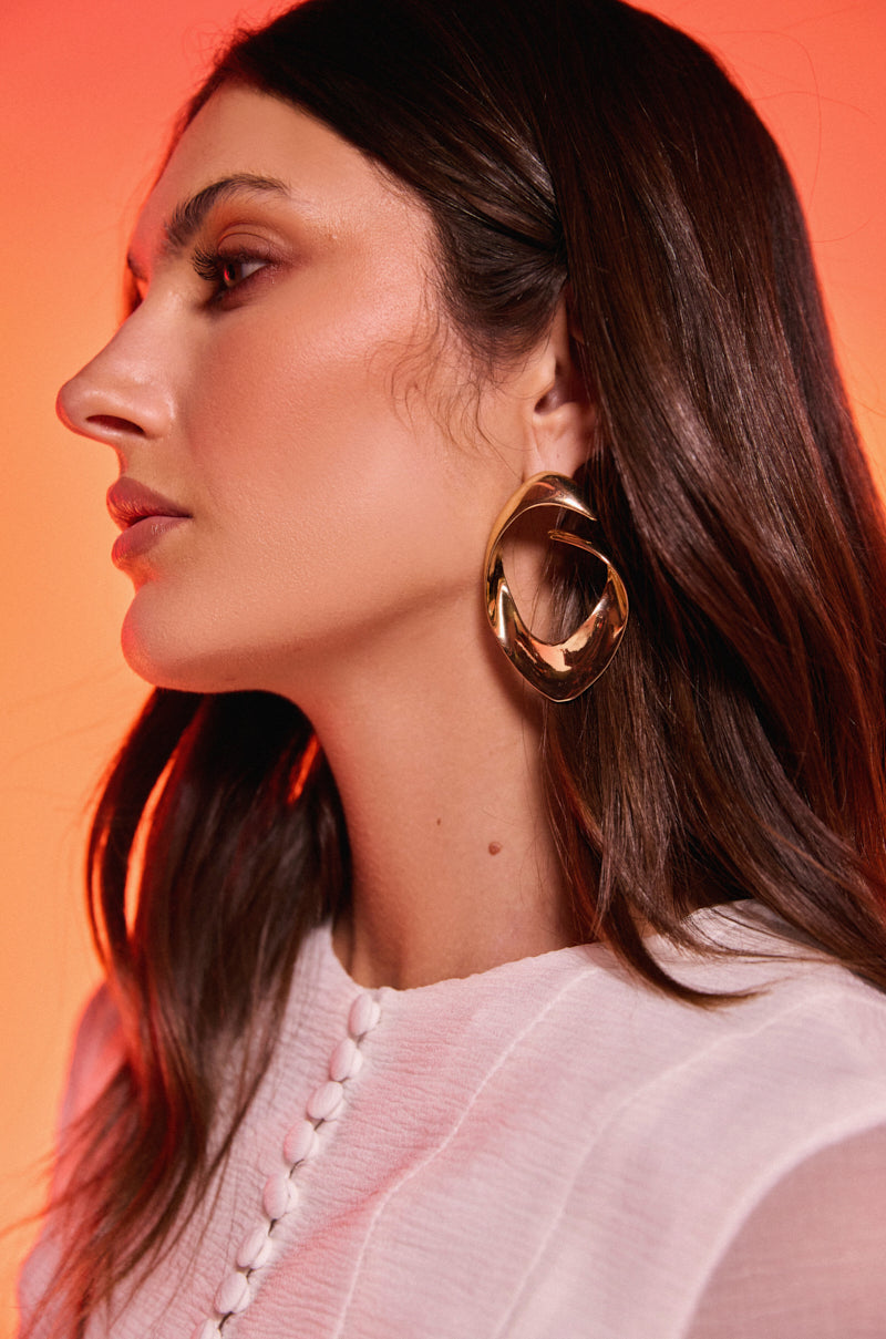 A woman with long brown hair, in a white textured top and the LOVERGIRL EARRING, poses in profile against a warm orange gradient background. 0