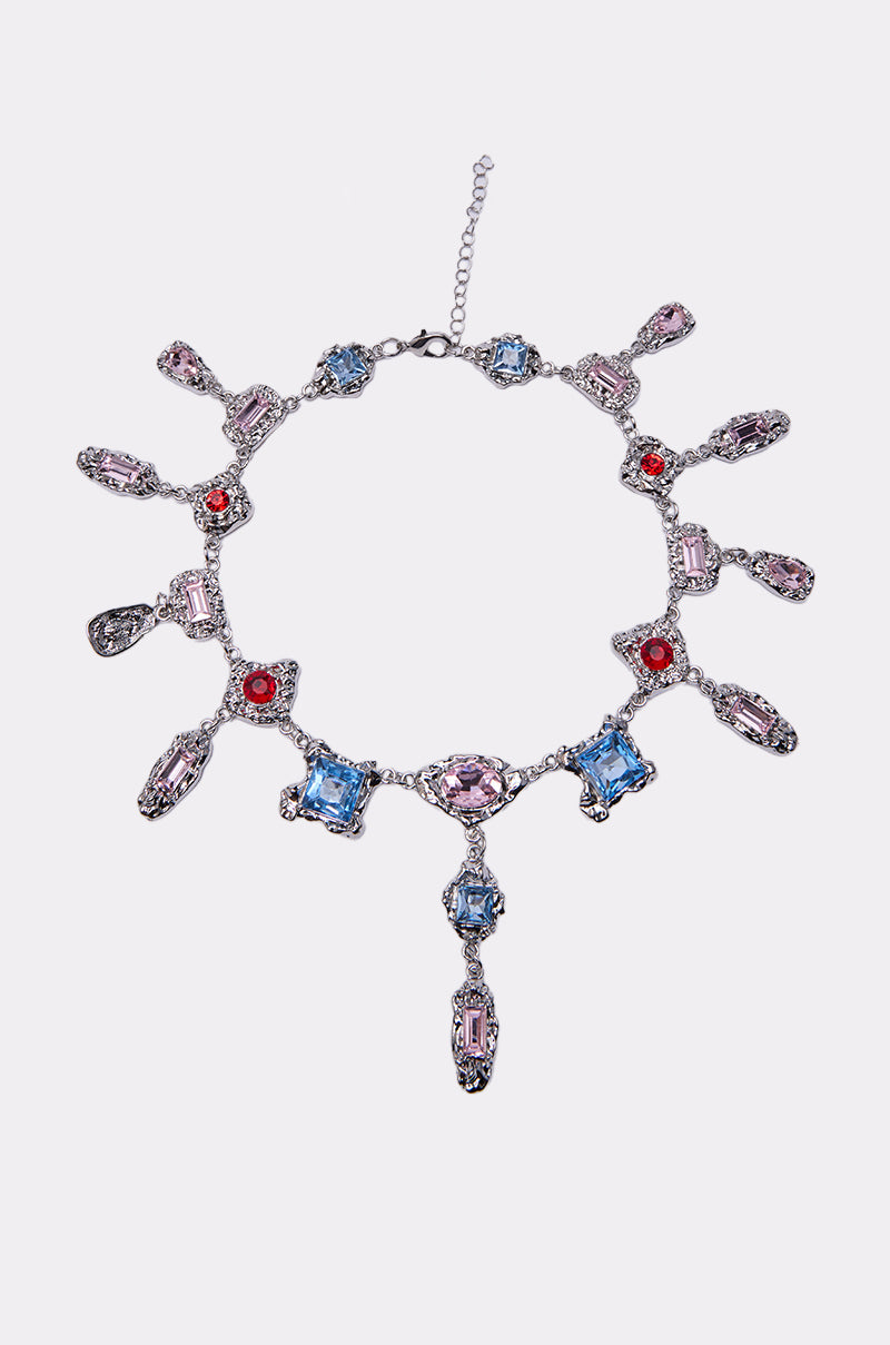 The EXOTIC LOVE NECKLACE is a silver piece adorned with blue, pink, and red gemstones in ornate settings, sparkling rhinestone accents, dangling gem drops, and includes an adjustable chain clasp.