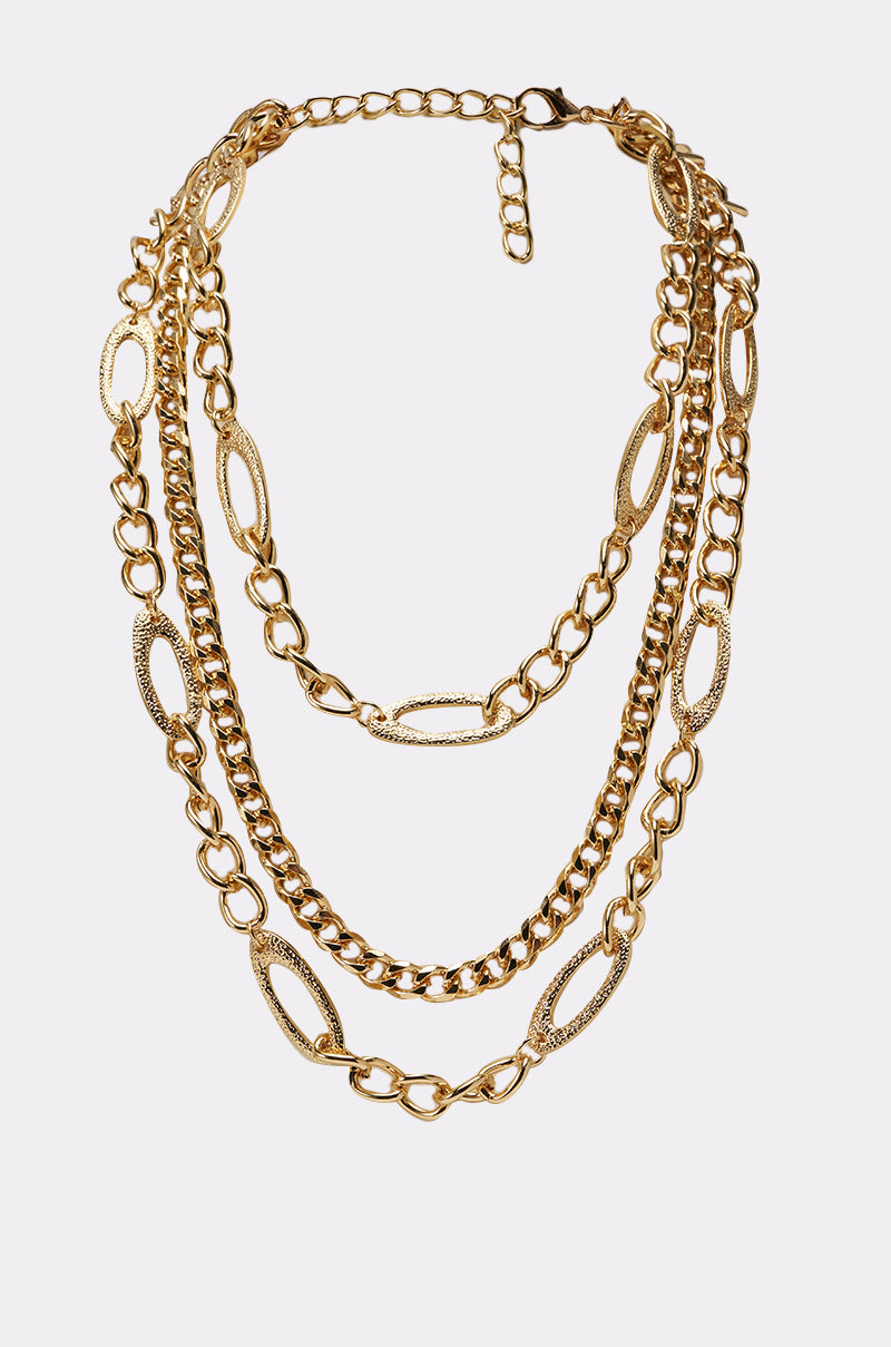 The LAYER ME UP NECKLACE is an adjustable layered gold chain necklace with three distinct chain styles, featuring oval and round links, shown against a plain white background. 1