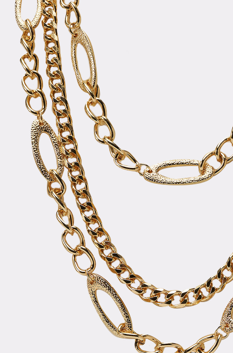 Close-up of the LAYER ME UP NECKLACE: one gold strand features elongated textured oval links, while the other has smaller curb-style links. Set against a light background, ideal for an adjustable layered look. 2