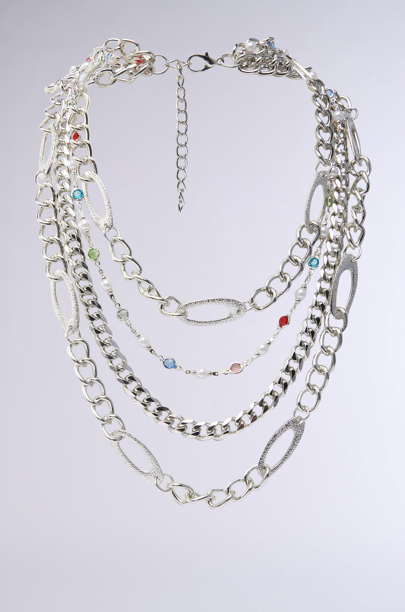 The TAKE A NOTE SILVER NECKLACE is a multi-layered piece with chunky silver chains in assorted link styles, accented by red, blue, and green gemstones, showcased on a light gray background.