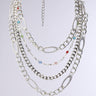 The TAKE A NOTE SILVER NECKLACE is a multi-layered piece with chunky silver chains in assorted link styles, accented by red, blue, and green gemstones, showcased on a light gray background.