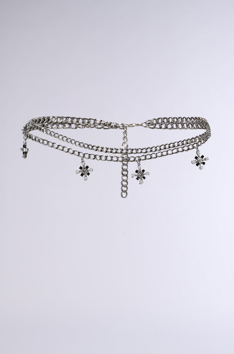 The DON'T CROSS ME BELT IN SILVER is a silver chain belt with a central hanging chain, four flower-shaped pendants, interlocking metal links, and floral designs. Adorned with gemstone embellishments, this elegant accessory adds sparkle to your festival look on a light gray background. 2