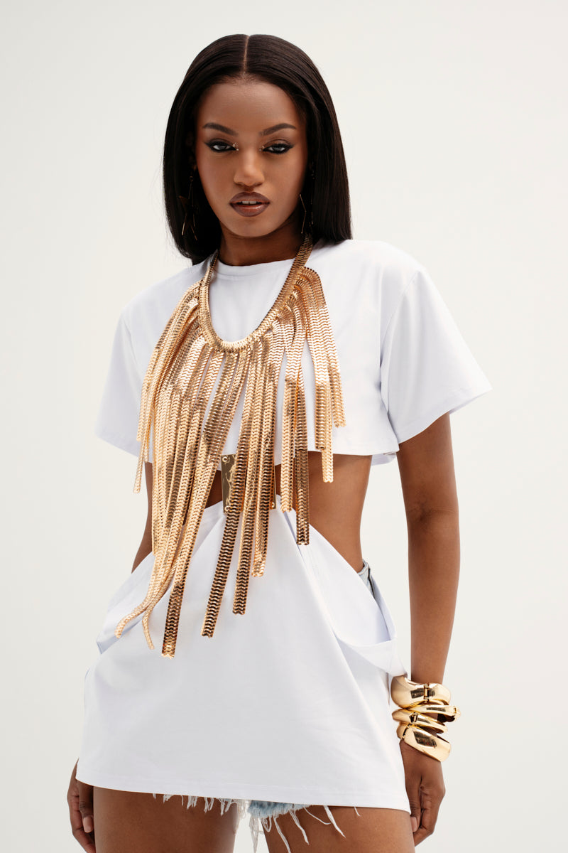 A stylish person wears a white cut-out top with the CANT TELL ME NO CHAIN NECKLACE, showcasing faux gold fringes. Their long, straight hair pairs well with denim shorts, while gold bangles accentuate their wrist against a plain background.
